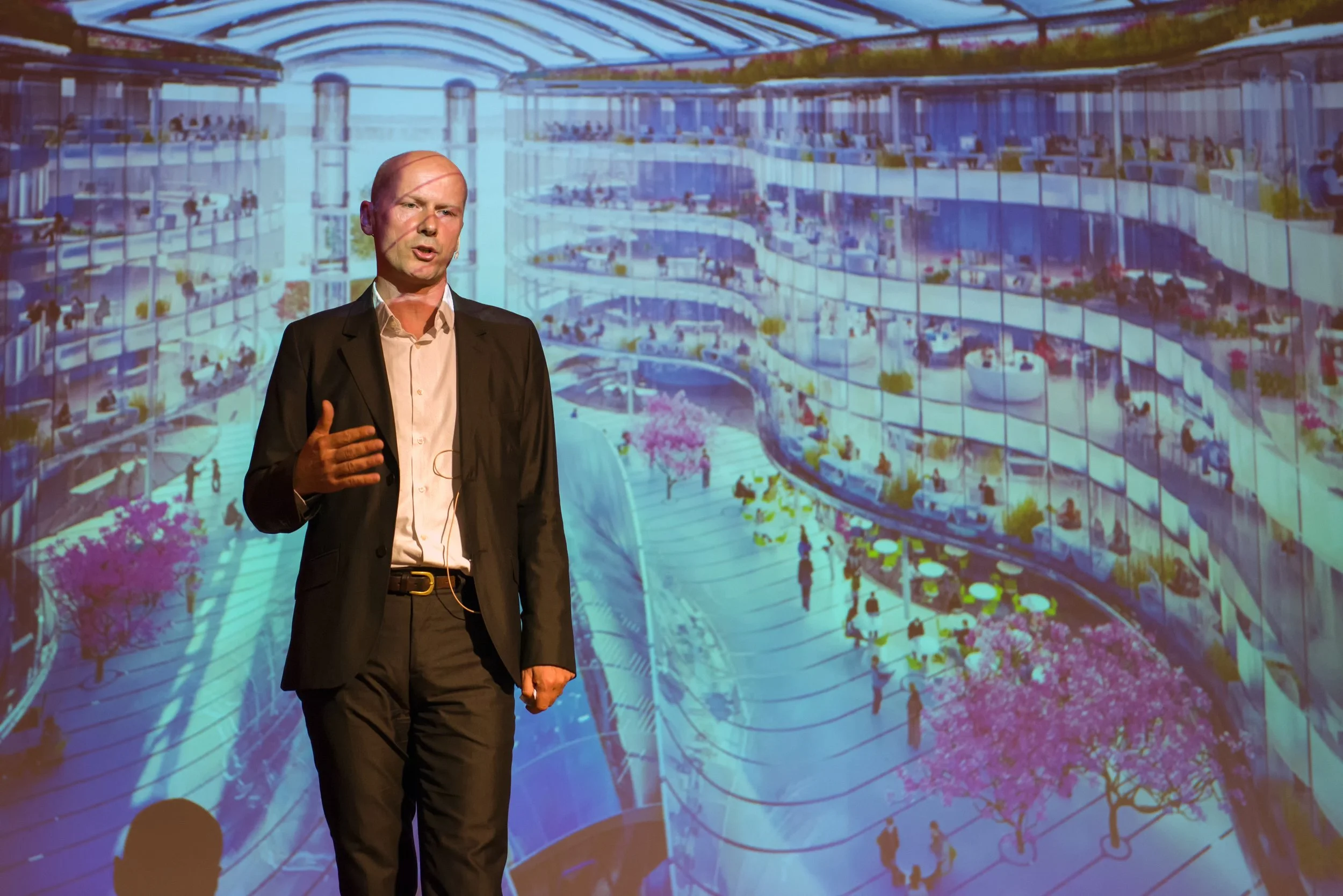 Man giving a presentation in front of a large digital rendering of a modern, multi-story building with a spacious indoor courtyard, glass walls, and several people inside and outdoors.
