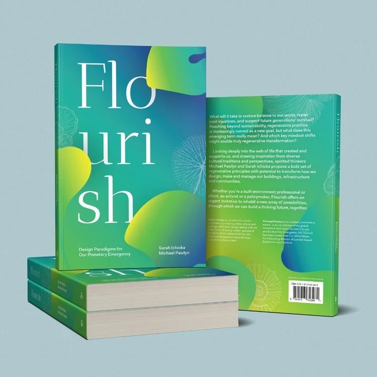 Three copies of a book titled 'Flourish' by Sarah Ichioka and Michael Pawlyn, with a blue and green cover, stacked, with one book open showing the back cover on a light gray surface.