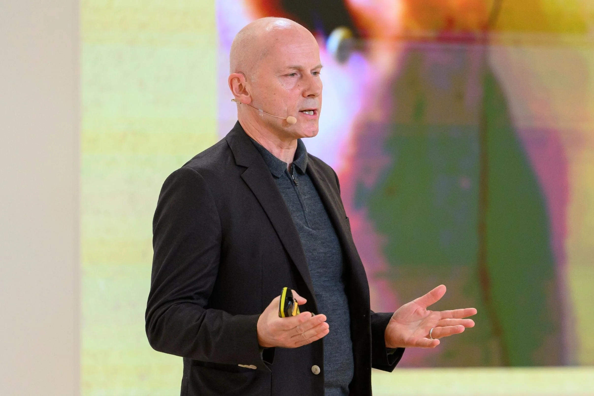 Michael Pawlyn wearing a black blazer and a dark gray shirt, speaking during a presentation, holding a yellow and black remote control, with a colorful background.
