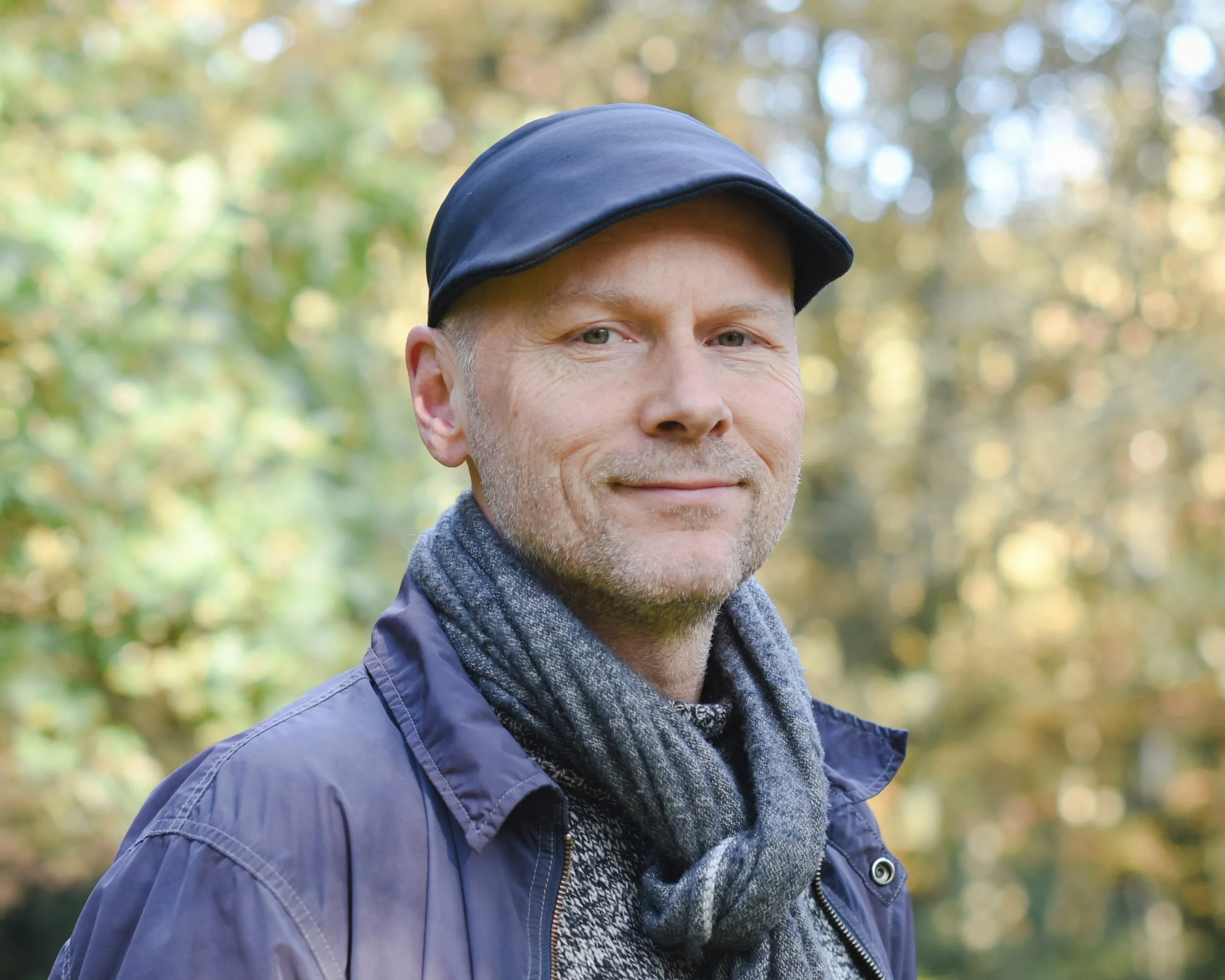 Michael Pawlyn, wearing a black cap, a gray scarf, and a blue jacket, standing outdoors with autumn foliage in the background.