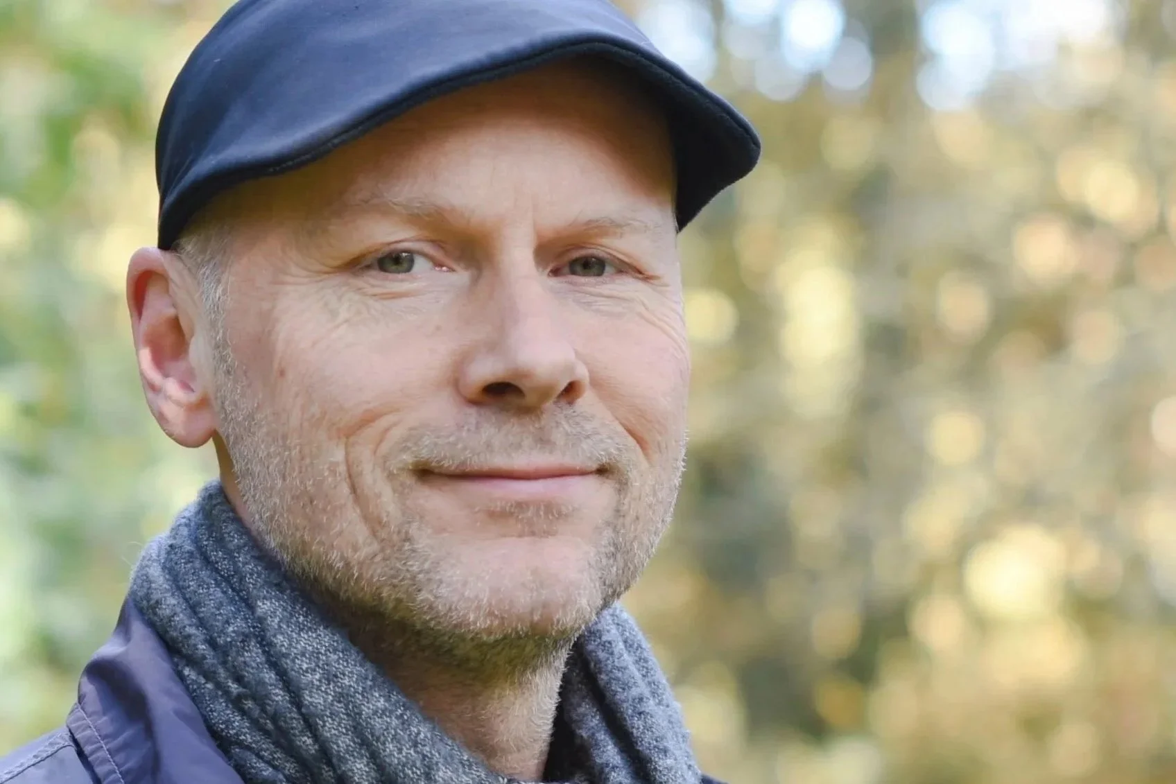 Close-up of a man outdoors, wearing a dark cap and a gray scarf, with a blurred background of trees and autumn leaves