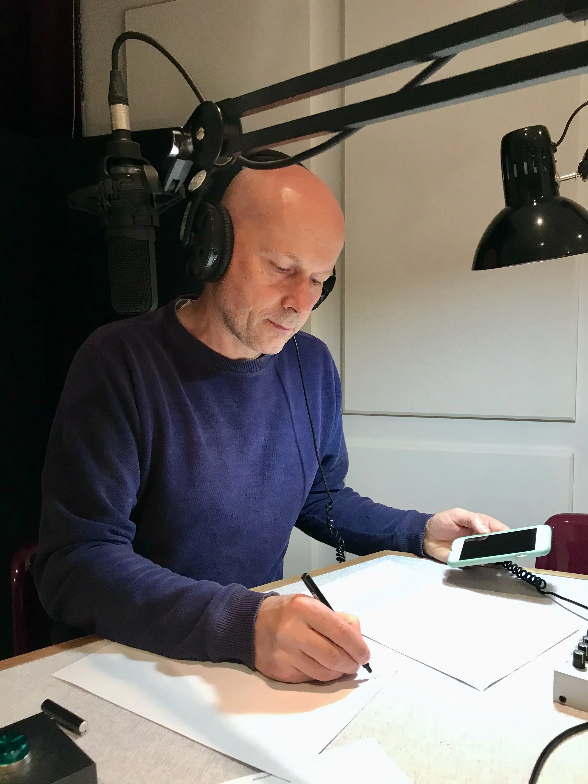 Michael Pawlyn wearing headphones is sitting at a desk, sketching on a piece of paper with a pen. He looks focused, with a smartphone mounted on a flexible stand in front of him. There are studio lights overhead.