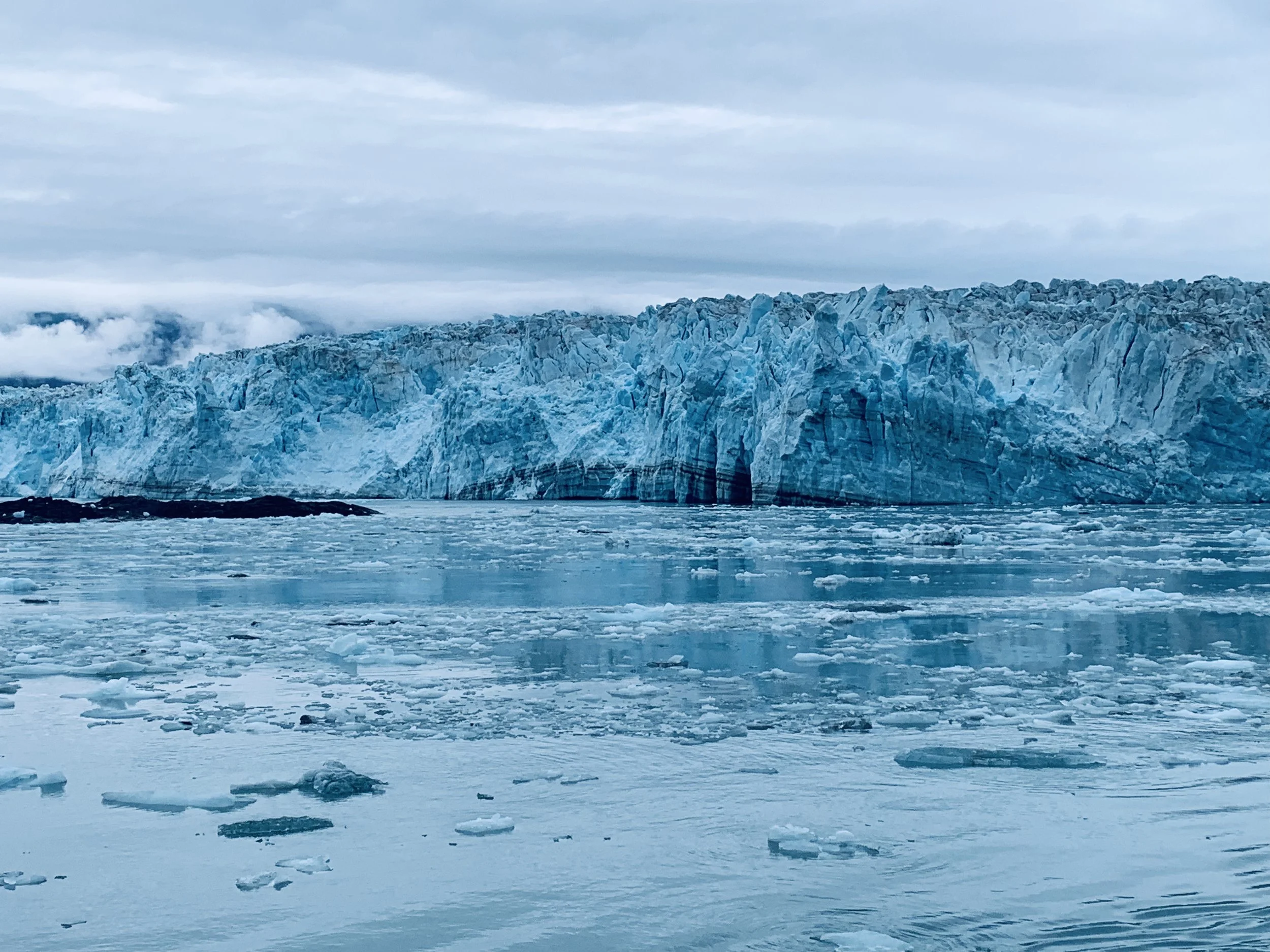 Massive Alaska tidewater glacier face with calved ice chunks floating in steel-blue water under overcast sky.