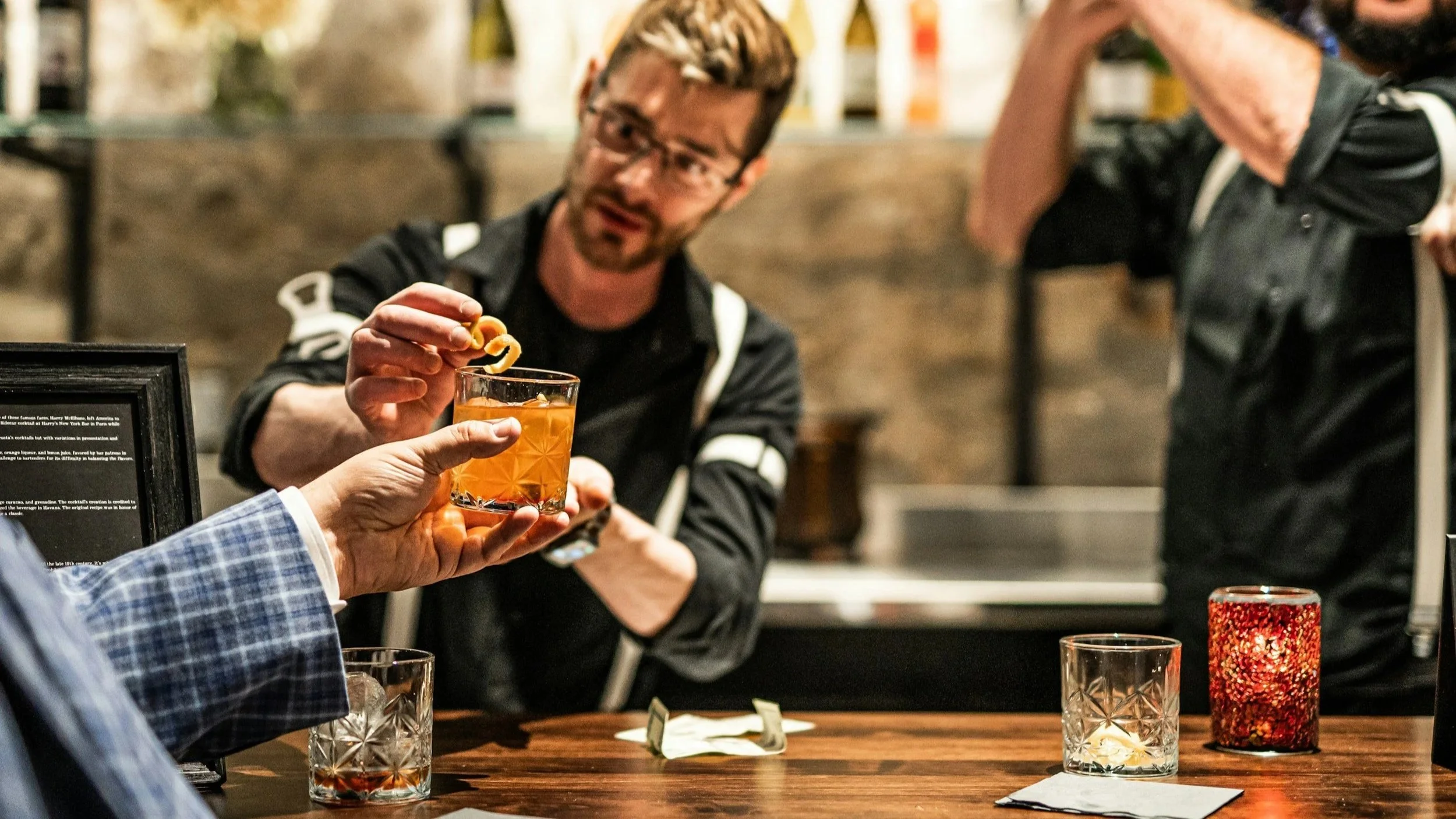 Bartender placing a garnish on a cocktail as a guest reaches for the glass in a warm, inviting bar setting.