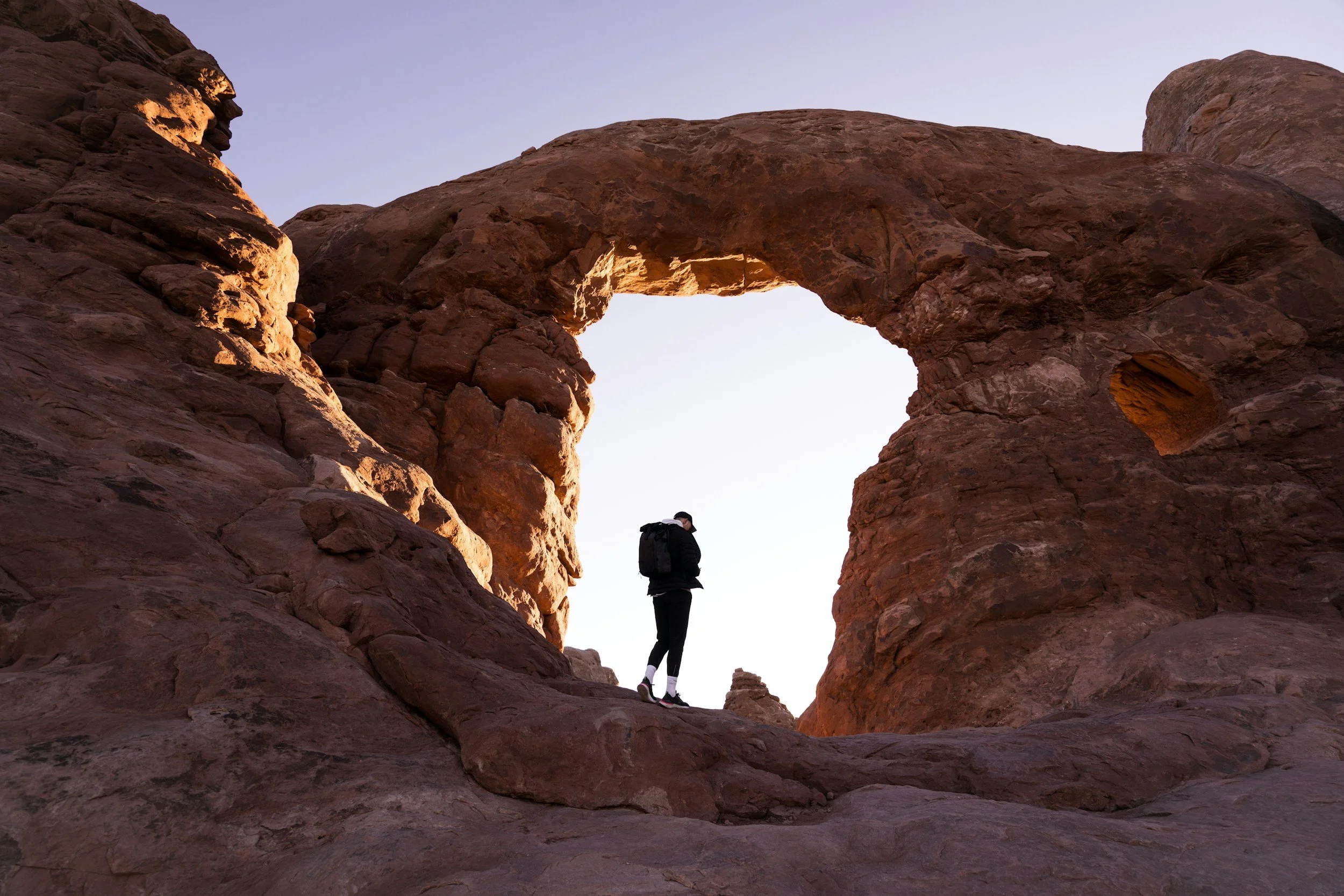 A person with a backpack standing on rocky terrain in front of a natural arch formation at sunset.