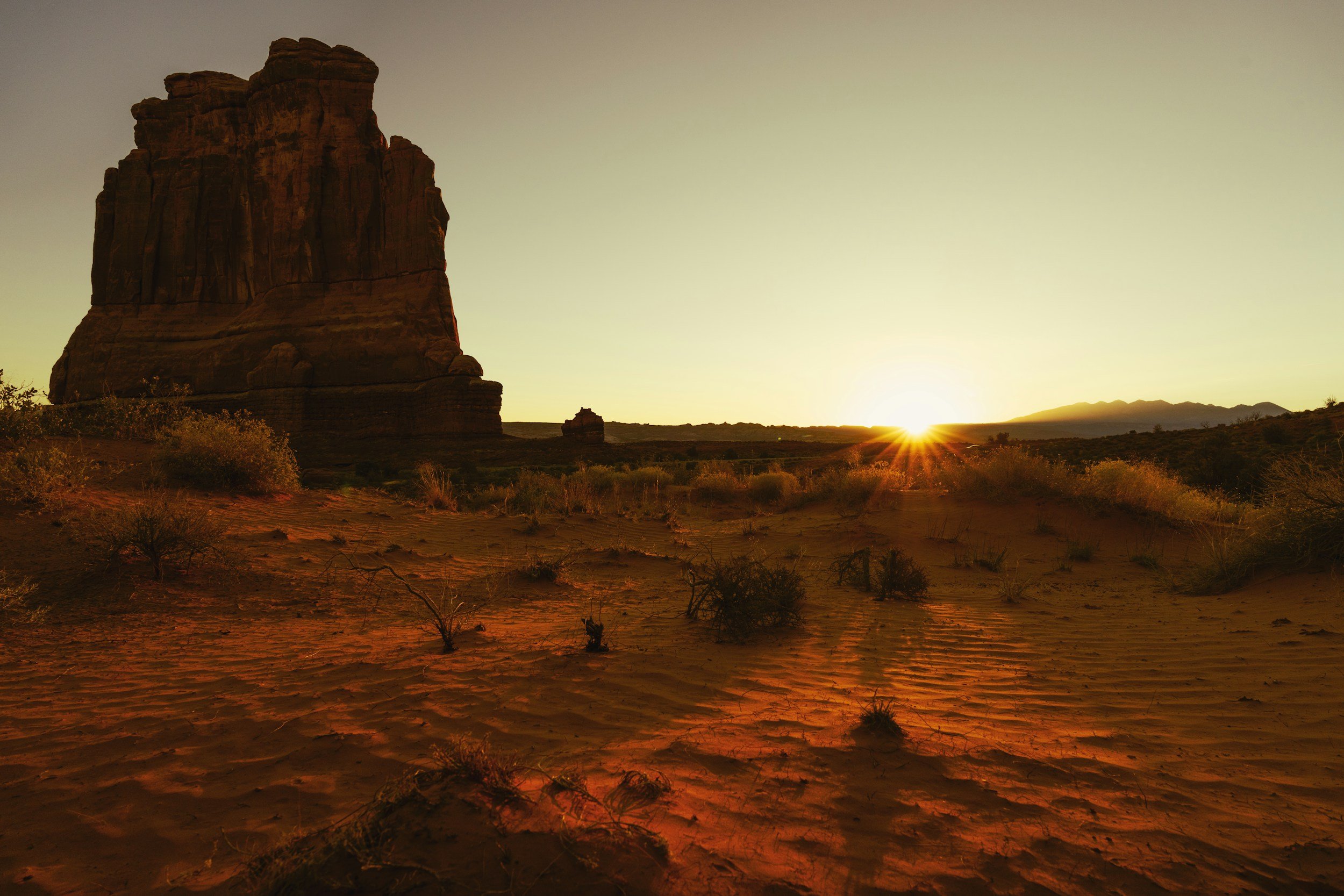 Red sandstone butte at Arizona desert sunrise with golden starburst light over rippled sand dunes, evoking the journey toward financial independence.