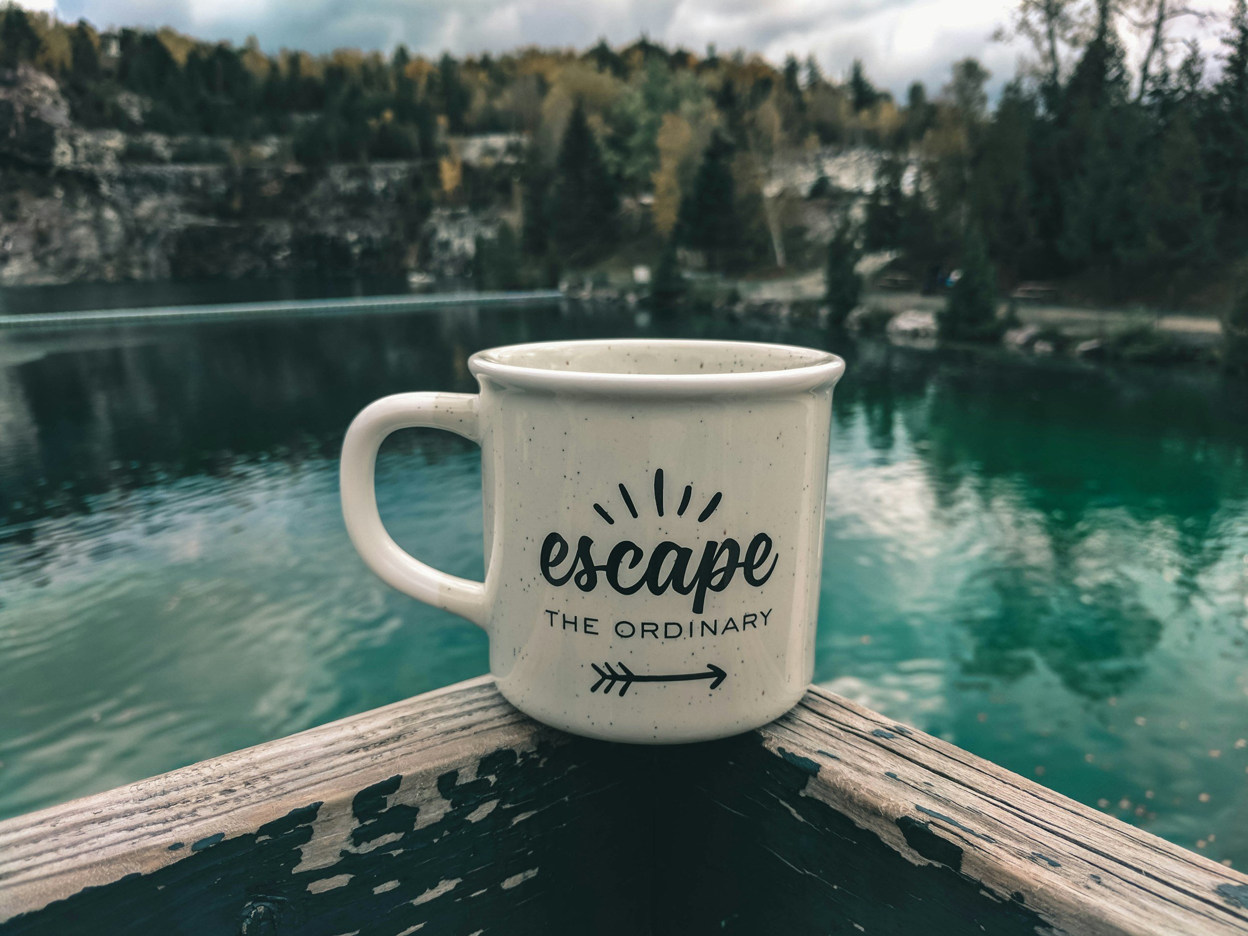 A ceramic mug reading "escape the ordinary" resting on a weathered railing overlooking a turquoise lake — the view from someone who timed the jump.