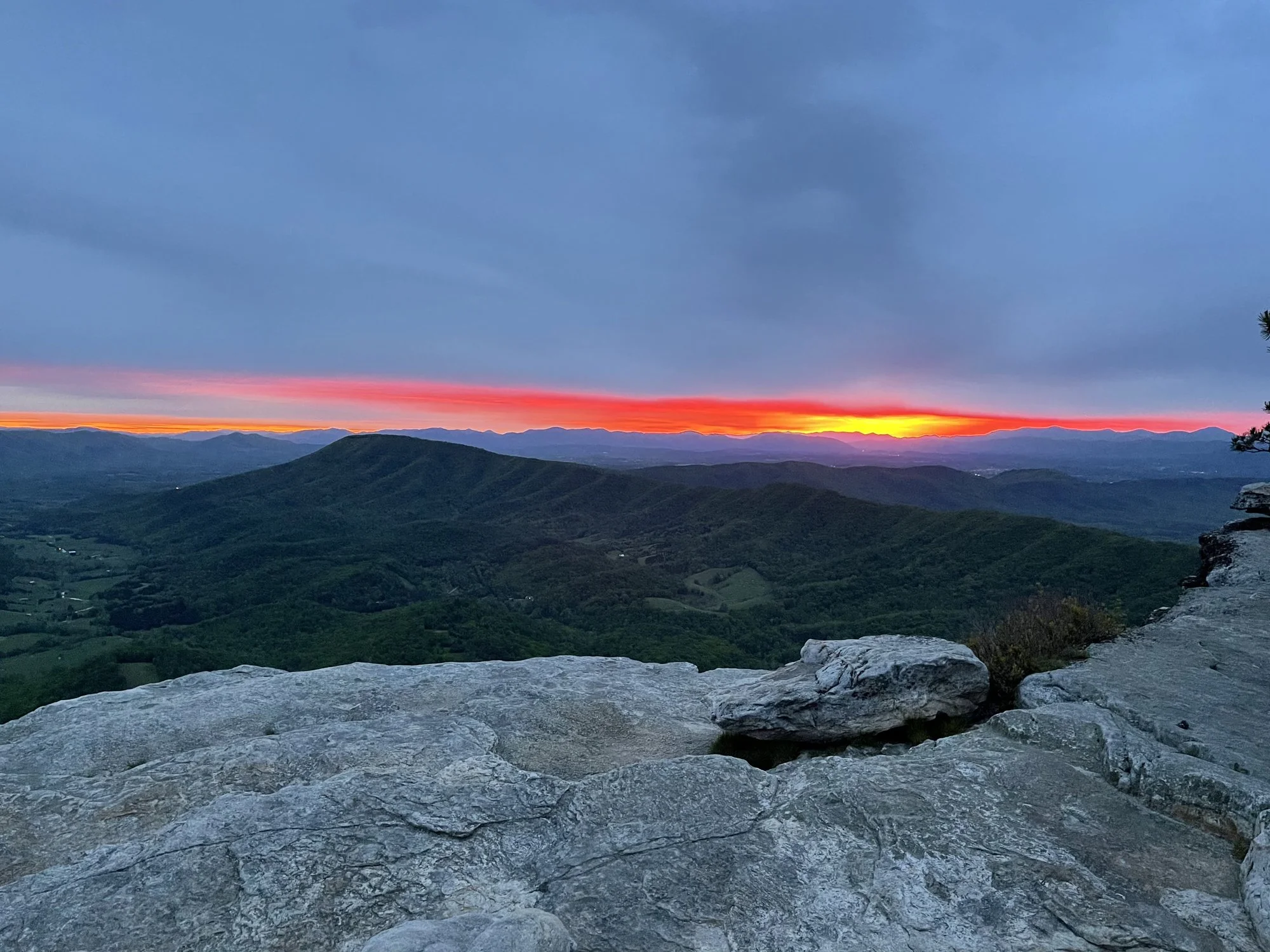Fiery red and orange sunset over Blue Ridge Mountain ridgeline viewed from rocky summit overlook, dark storm clouds above.