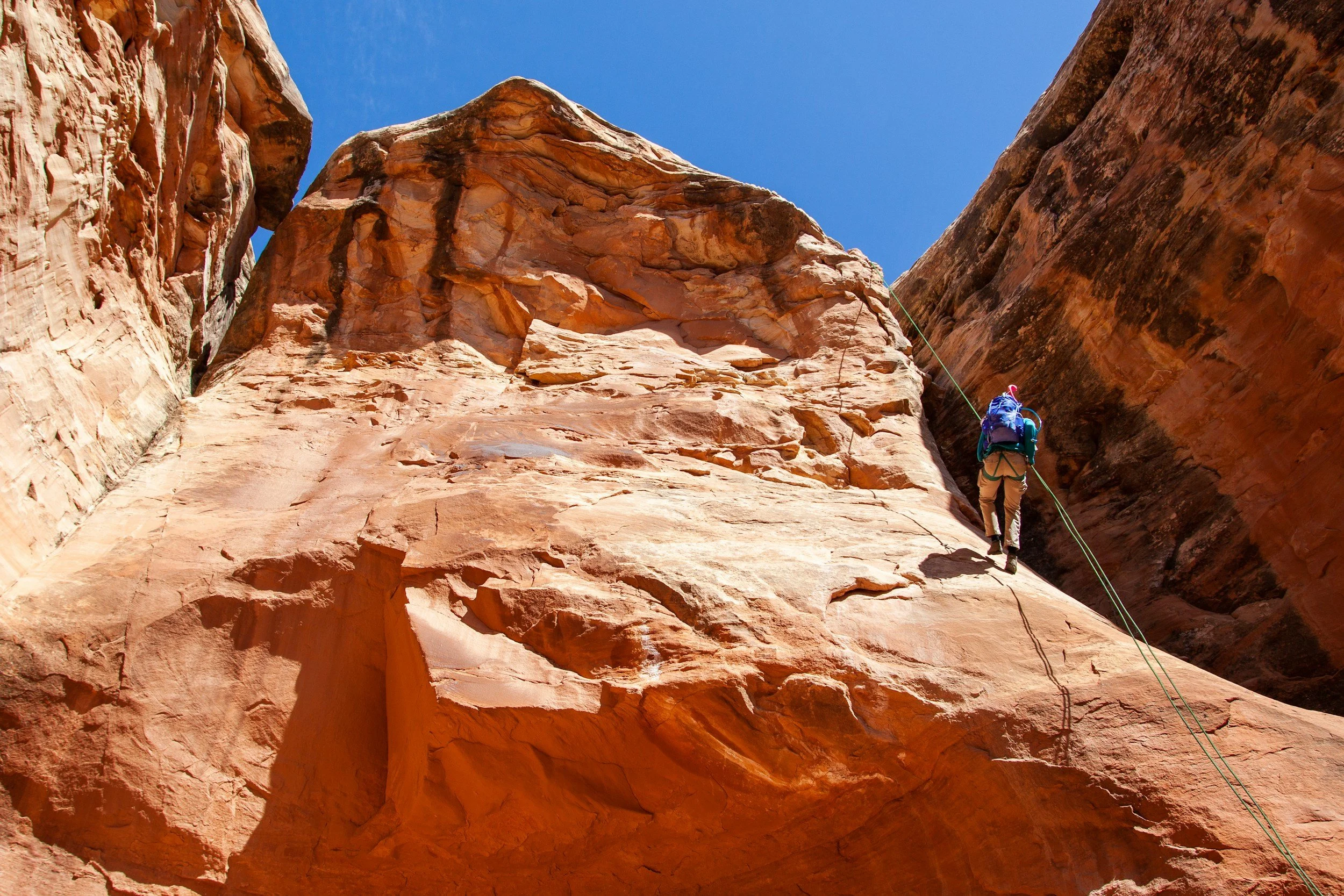 Hiker with blue backpack rappelling down sheer red sandstone cliff face in a narrow desert canyon, green rope visible against warm orange rock, blue sky visible at top between towering canyon walls.