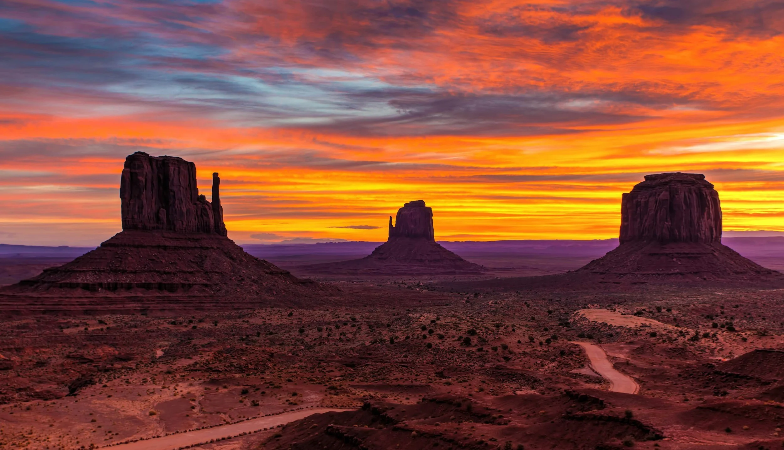 A dirt road winds between the iconic sandstone buttes of Monument Valley at sunrise, the sky blazing terracotta and gold above the desert floor. Two paths, one choice, and the American Southwest as witness.