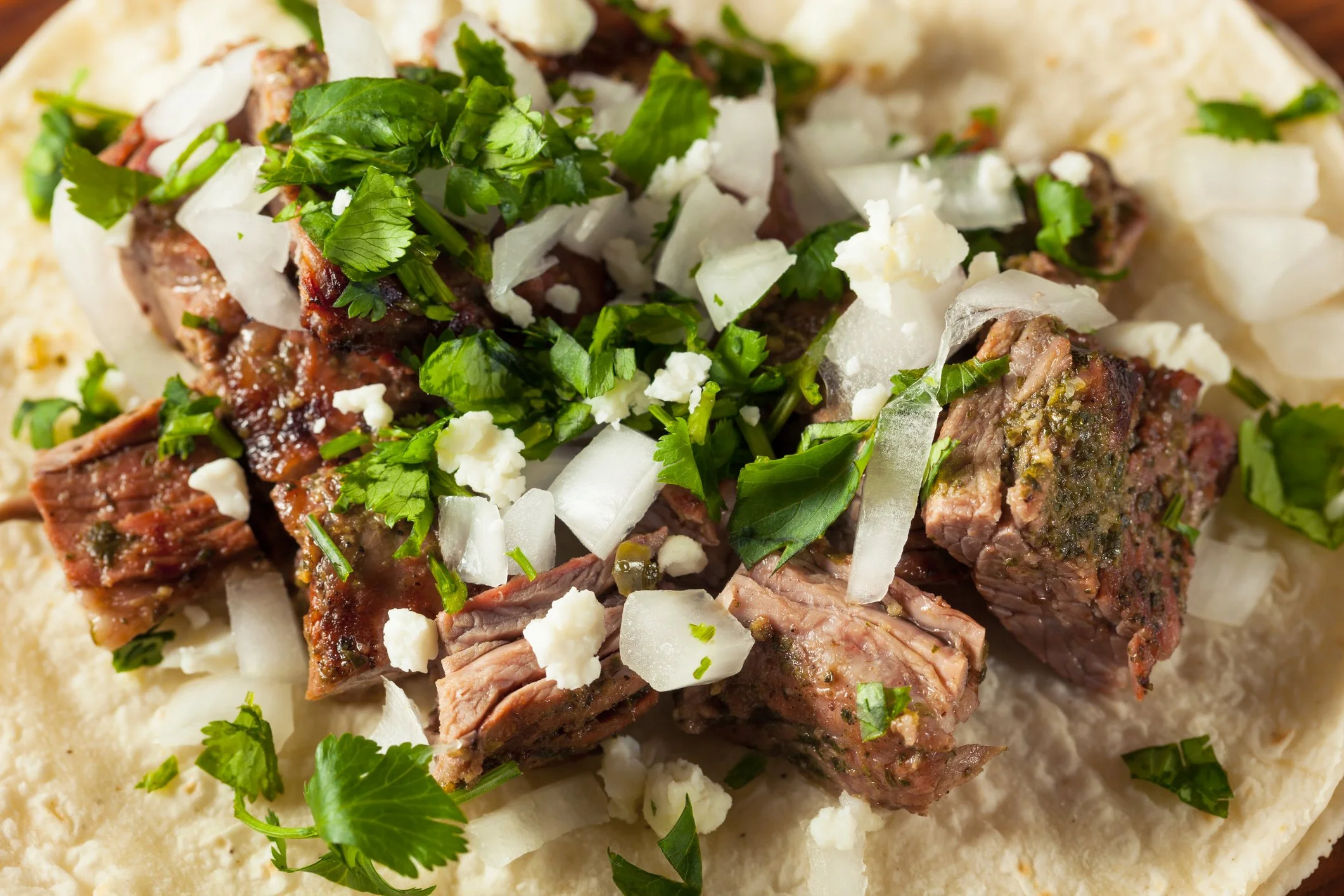 Close-up of authentic carne asada street taco on flour tortilla topped with fresh cilantro, diced white onion, and crumbled cotija cheese.