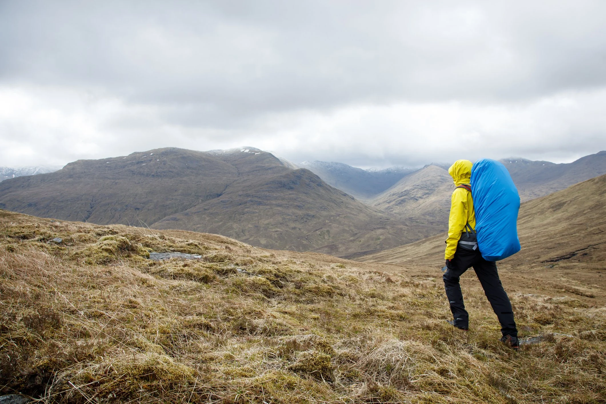 Solo hiker in a yellow rain jacket with a blue pack cover walking through windswept mountain grass in overcast conditions with mountains disappearing into clouds.