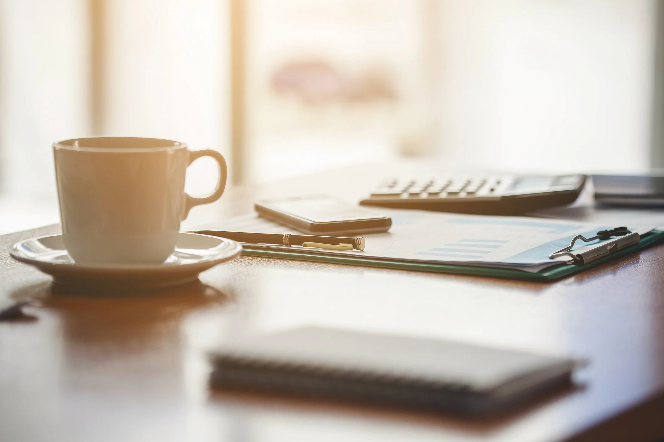 A coffee cup, calculator, and financial documents on a wooden table in warm morning light, reflecting the quiet Saturday morning work of building a retirement budget.