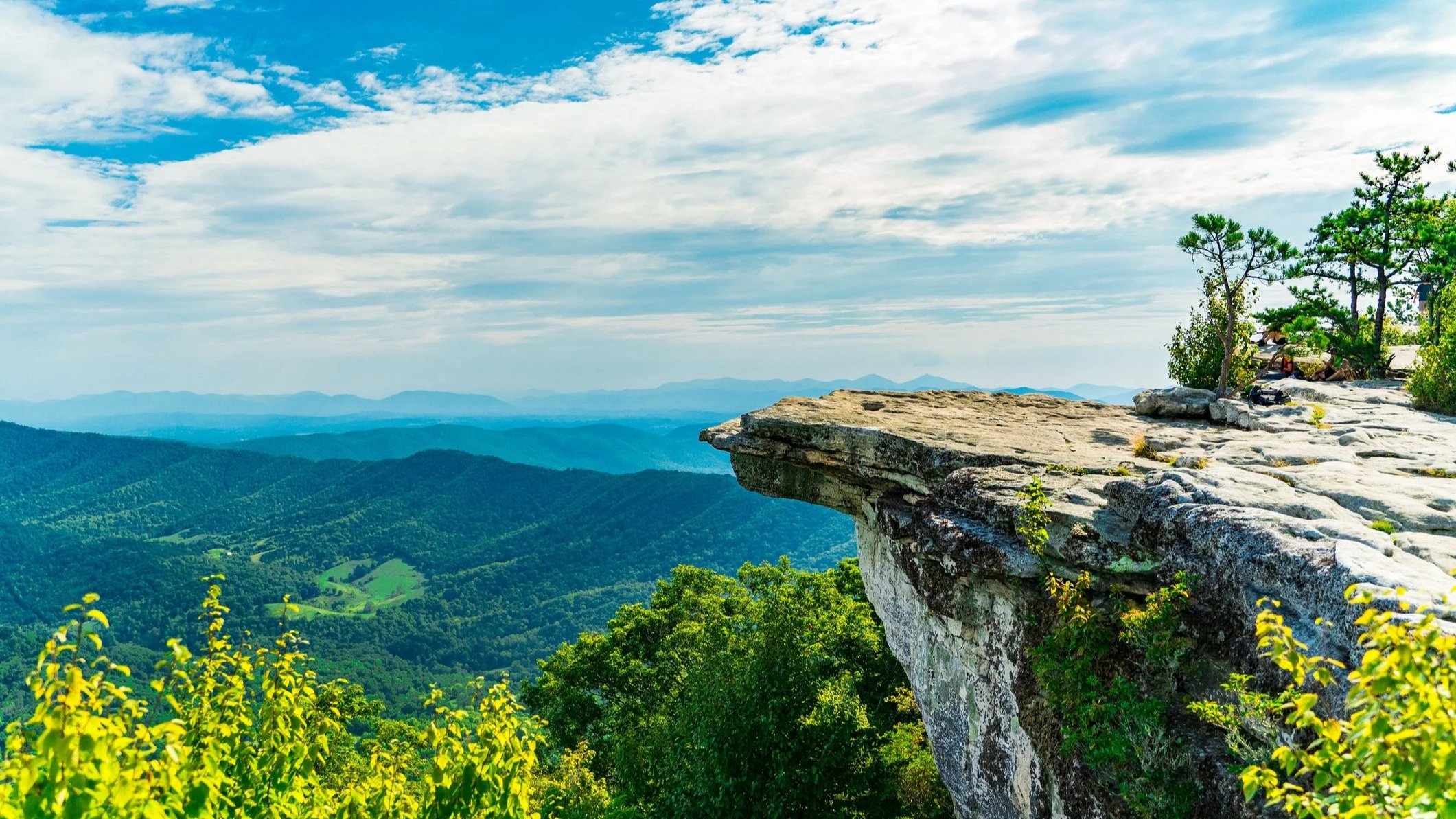 Stone rock ledge jutting over lush Blue Ridge Mountain valley, hikers resting at summit overlook under partly cloudy sky.