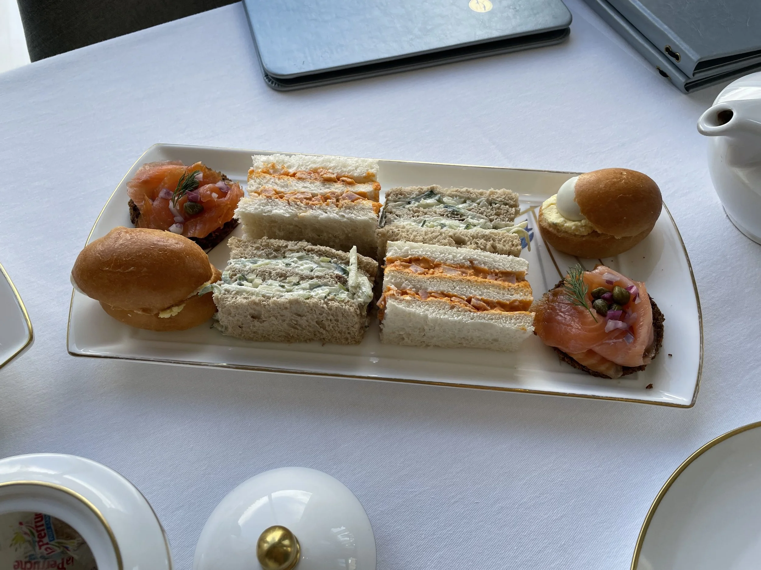 Elegant white porcelain tray of traditional high tea finger sandwiches, gold-rimmed teacups, and teapot on white linen tablecloth.