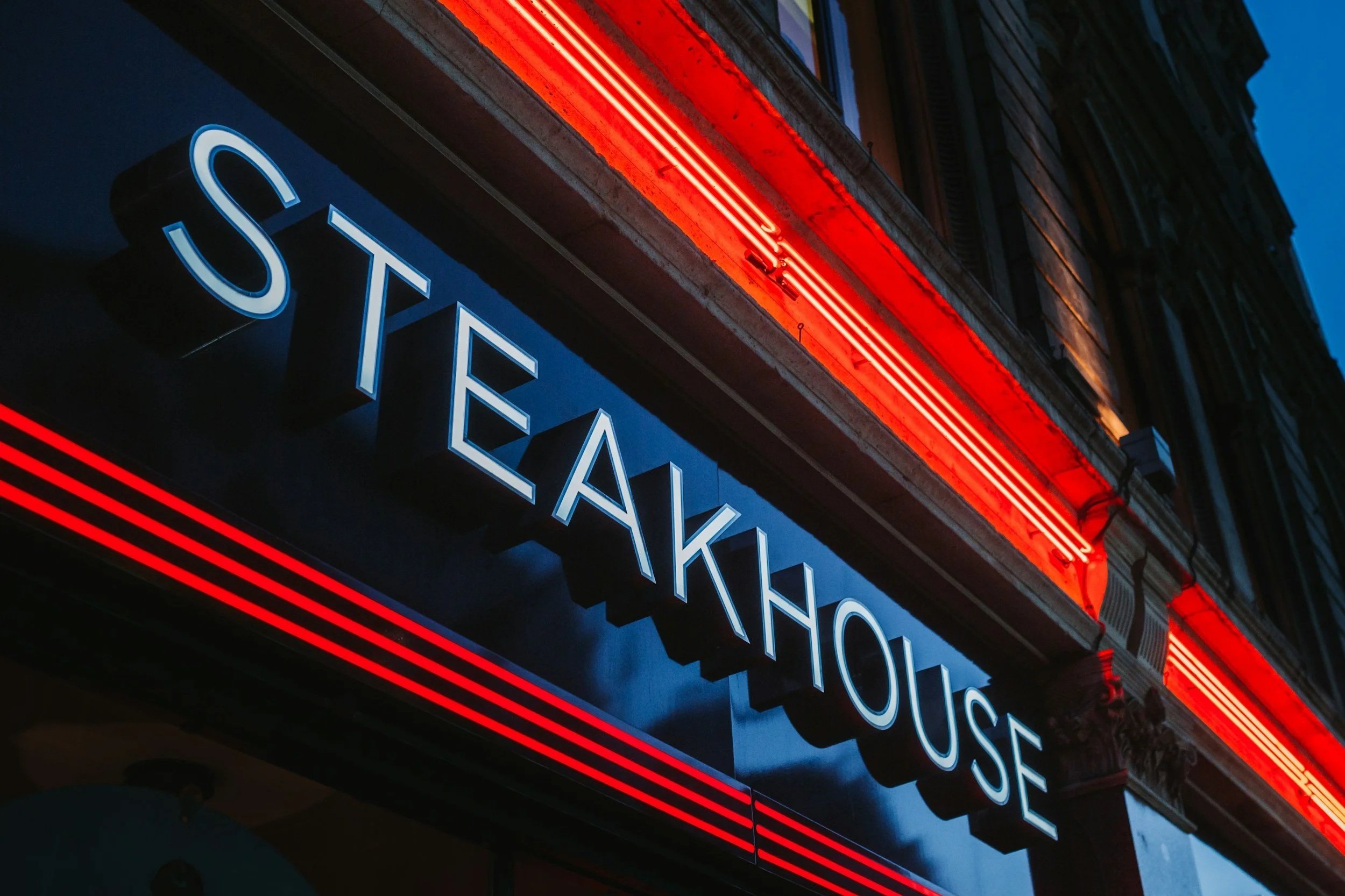Steakhouse exterior with glowing neon sign at dusk, evoking the world of financial abundance and the first threshold of enough.