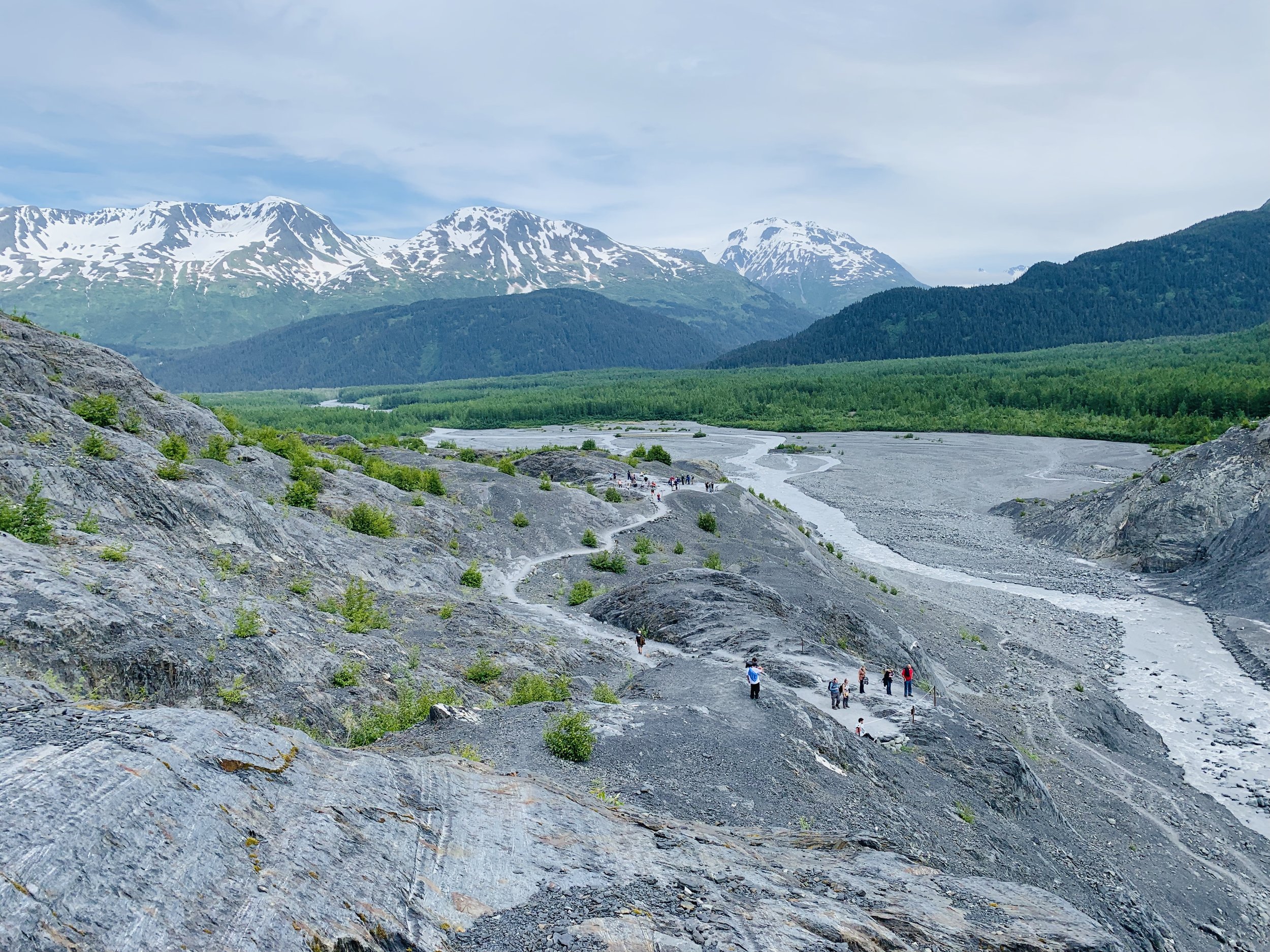 Hikers exploring rocky glacial moraine valley in Alaska with braided river, dense forest, and snow-capped mountain peaks under partly cloudy sky.