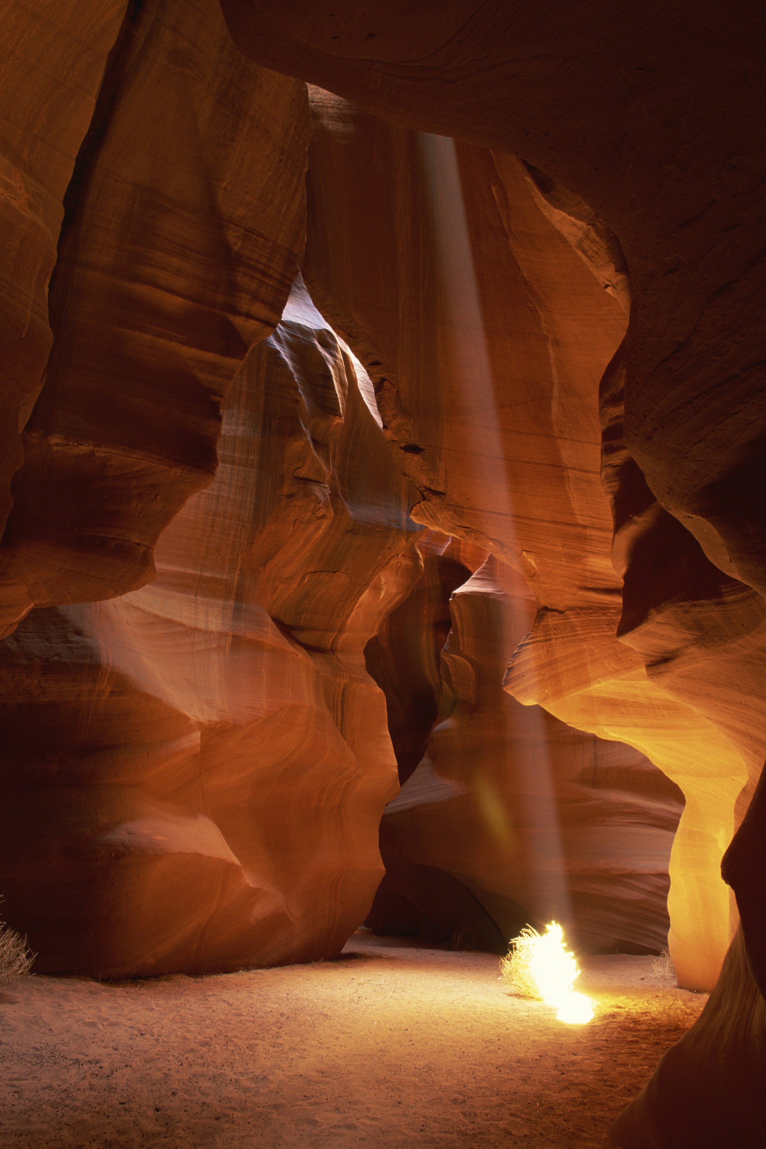 Inside a narrow slot canyon with smooth, wavy rock walls in reddish-brown and orange tones. A vertical light beam shines down, illuminating the sandy floor and creating a glow near the base.