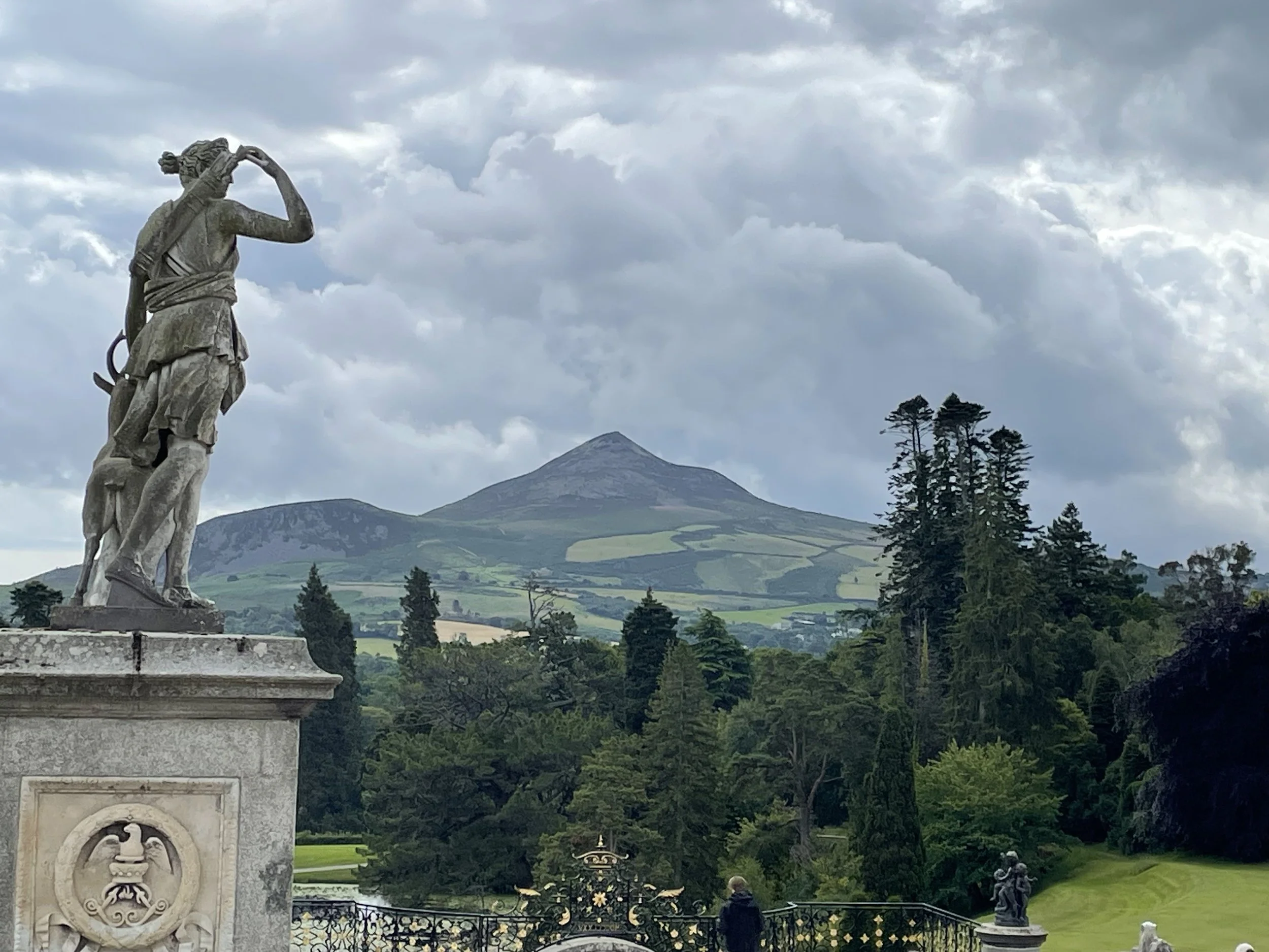 Classical stone statue on ornate pedestal overlooking Irish estate gardens with tall evergreens, lush green hillside farmland, and dramatic conical mountain peak under stormy cloudy sky.
