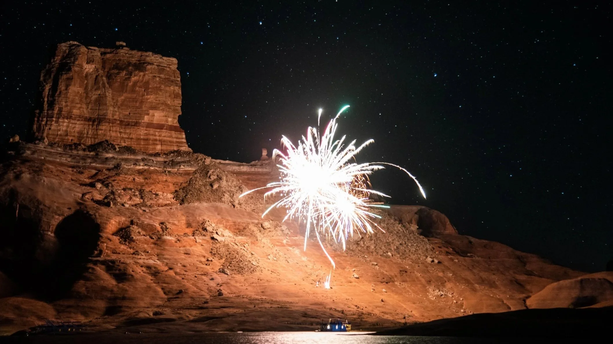 Brilliant white firework exploding against a star-filled night sky beside a towering red sandstone butte at Lake Powell, small houseboat illuminated on dark water below.