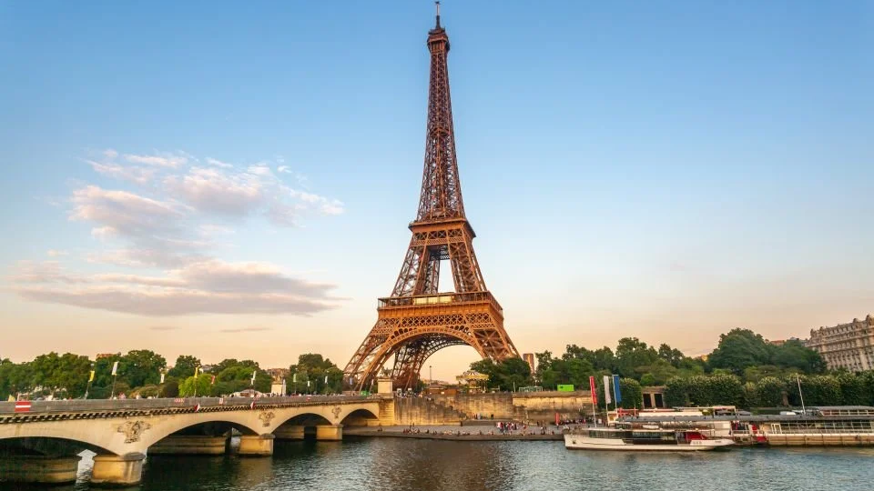 Eiffel Tower rising above the Seine River at golden hour, stone bridge in foreground, riverboat docked along tree-lined Paris embankment under soft evening sky.