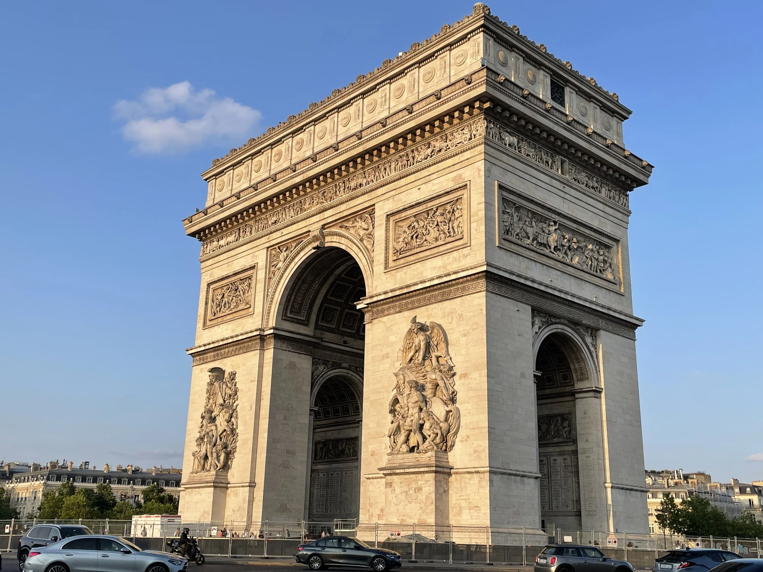 Arc de Triomphe in Paris bathed in warm golden evening light, intricate stone relief sculptures on facade, traffic and buildings visible at base under clear blue sky.