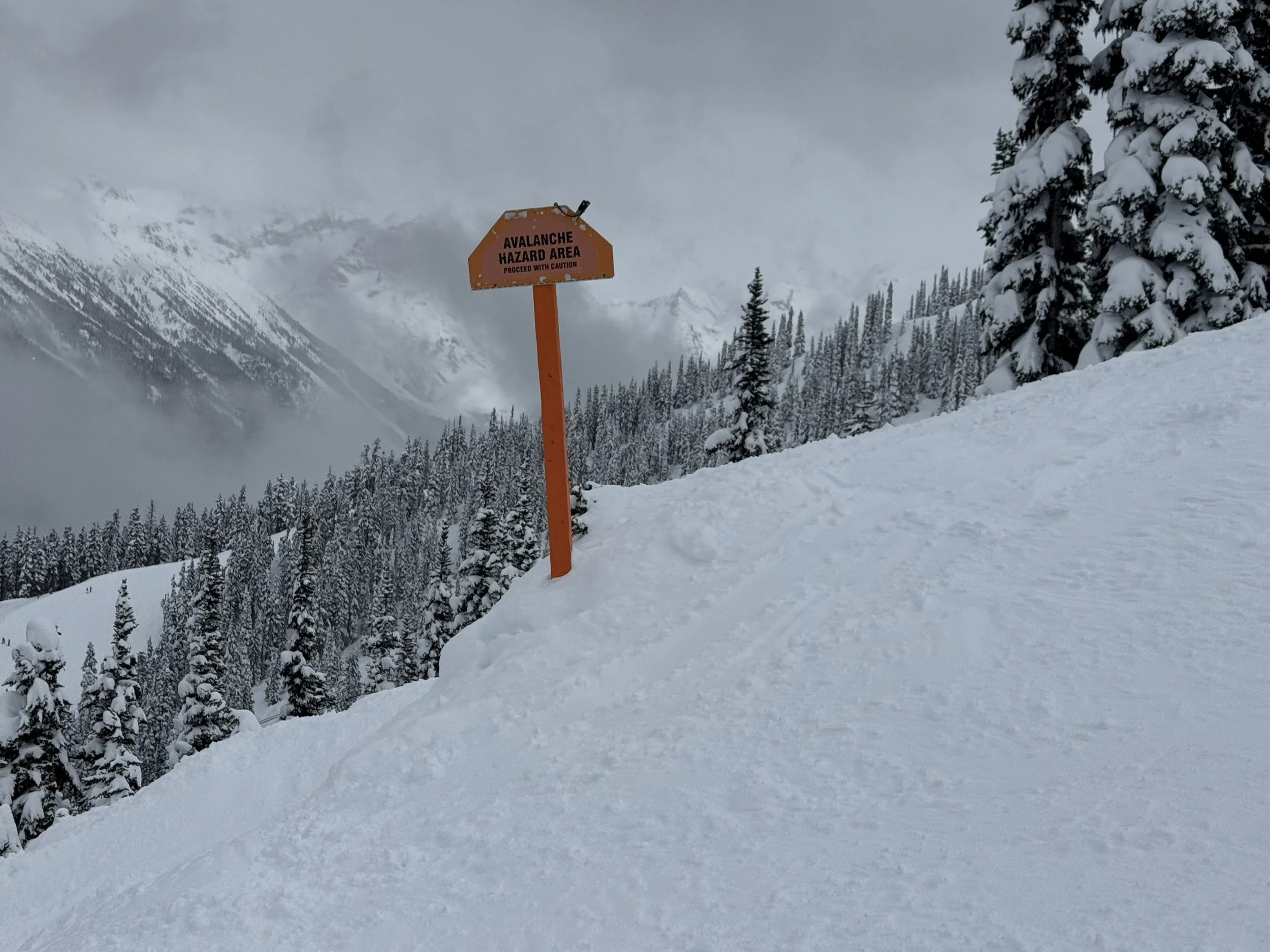 Orange avalanche hazard warning sign on a steep snow-covered slope at Whistler Blackcomb, with cloud-shrouded mountain peaks and forested valley visible in the background.