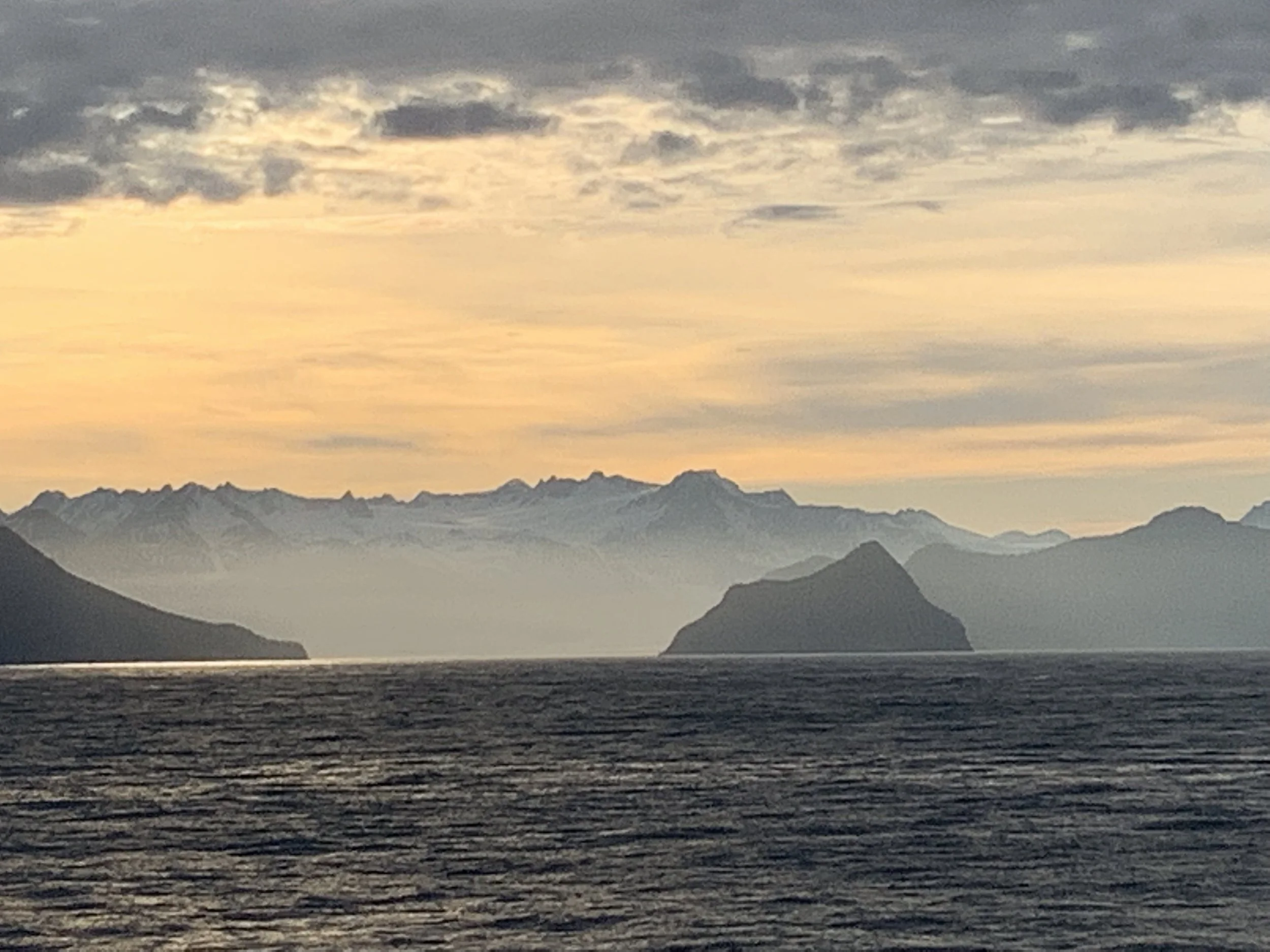 Golden sunset over calm Alaska Inside Passage waters with layered silhouetted mountain ranges and glaciers fading into misty horizon.