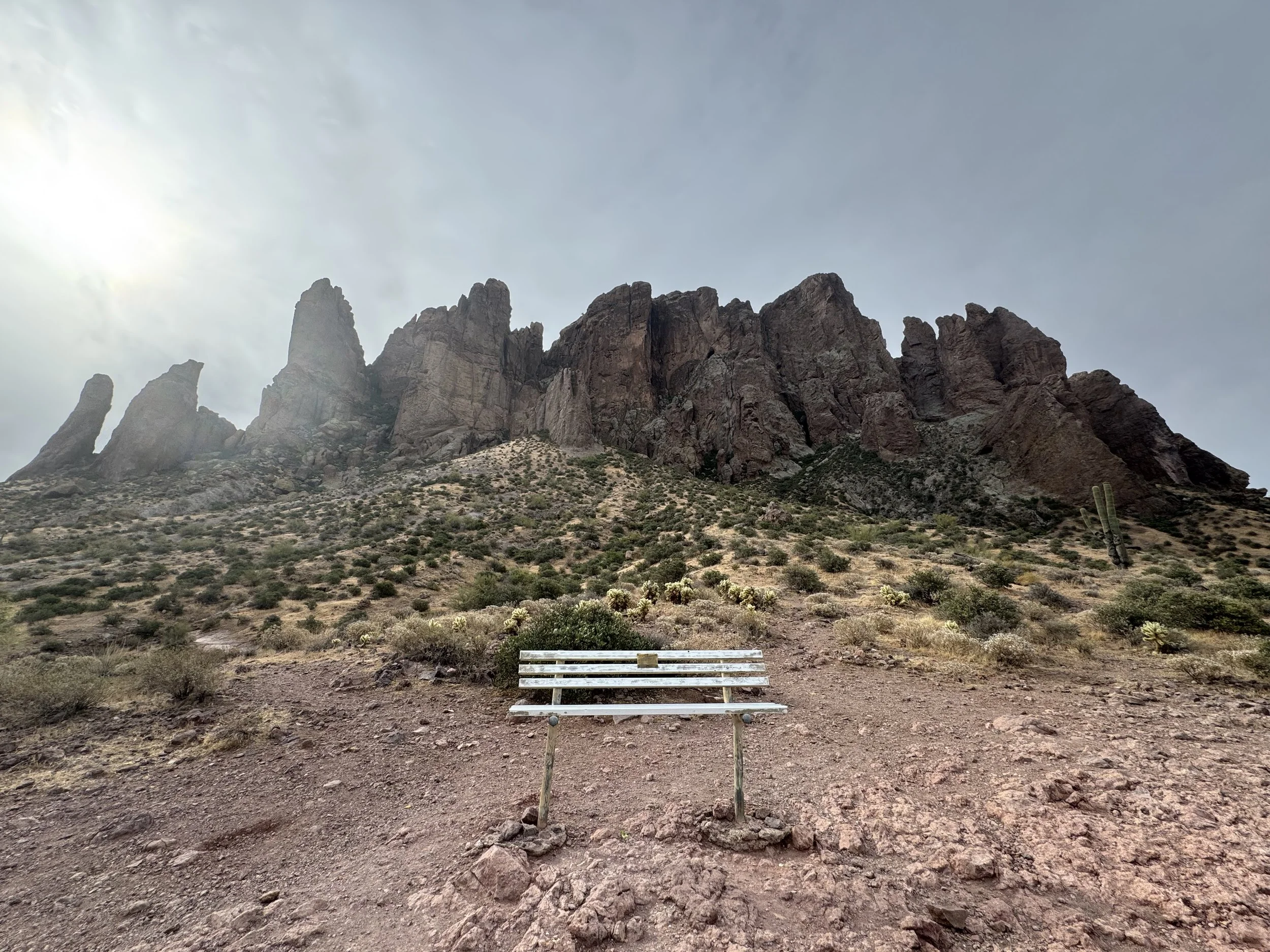 An empty white bench on an Arizona desert trail below dramatic rock spires on an overcast morning — a place to think through the harder questions.