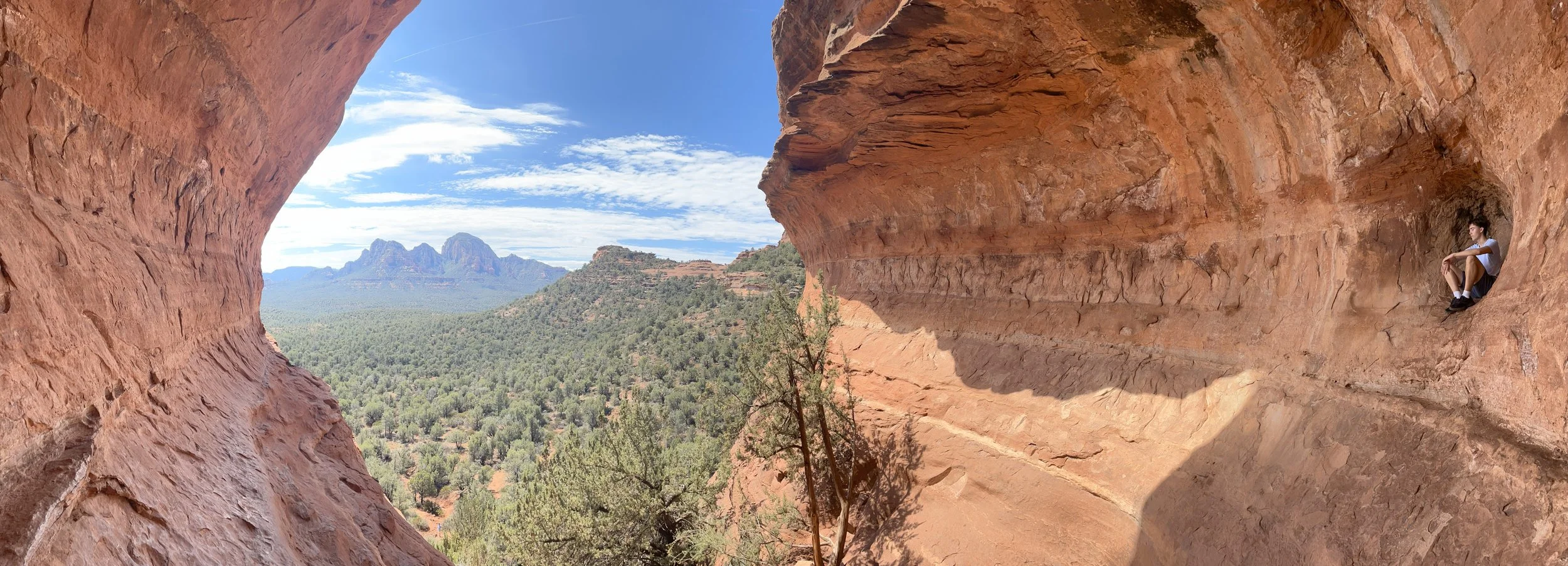 A figure resting small inside a Sedona cave opening, looking out toward a vast landscape of red rock and forest — sheltered by the old, facing the new.