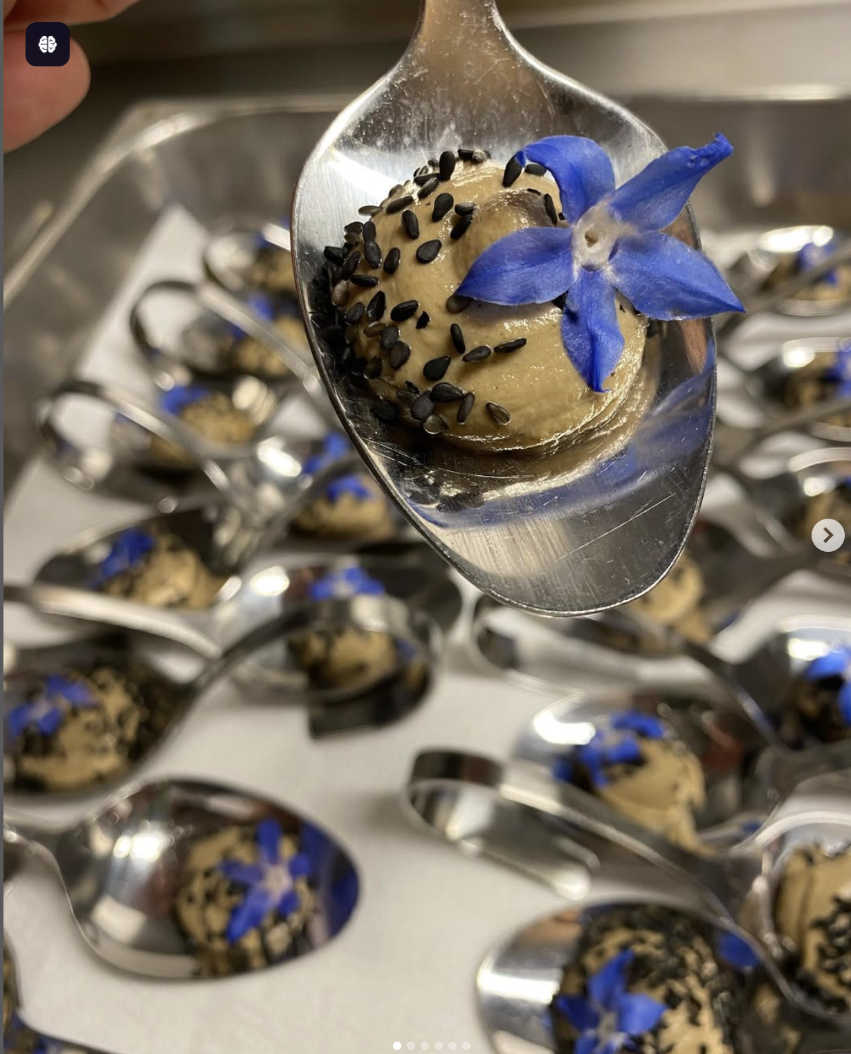 Häppchen mit einer blauen Blume und schwarzen Saaten auf einem Löffel, mehrere ähnliche Häppchen auf Löffeln im Hintergrund.