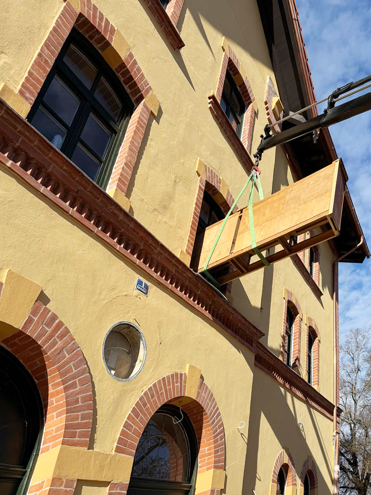Ein Baugerüst mit einem Holzsockel vor einem gelben Gebäude mit roten Ziegelbögen um die Fenster, im Vordergrund Teile der Gebäude, im Hintergrund blauer Himmel mit Wolken.