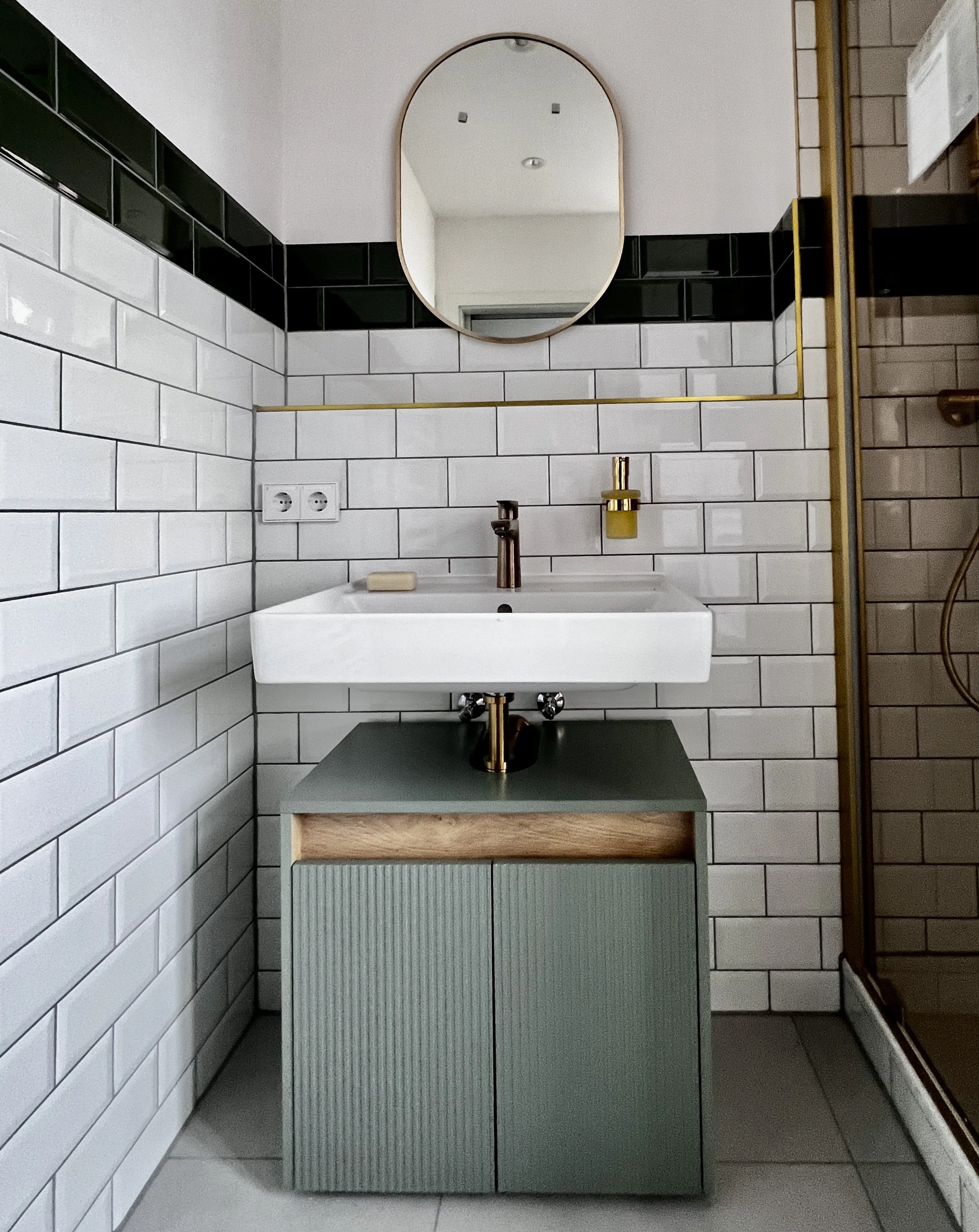 Ein moderner Badezimmerwaschbecken mit Spiegel und grauer Unterschränke, weiße Wandfliesen mit schwarzen Fliesen an der oberen Wand, goldene Accessoires und Dusche sichtbar.