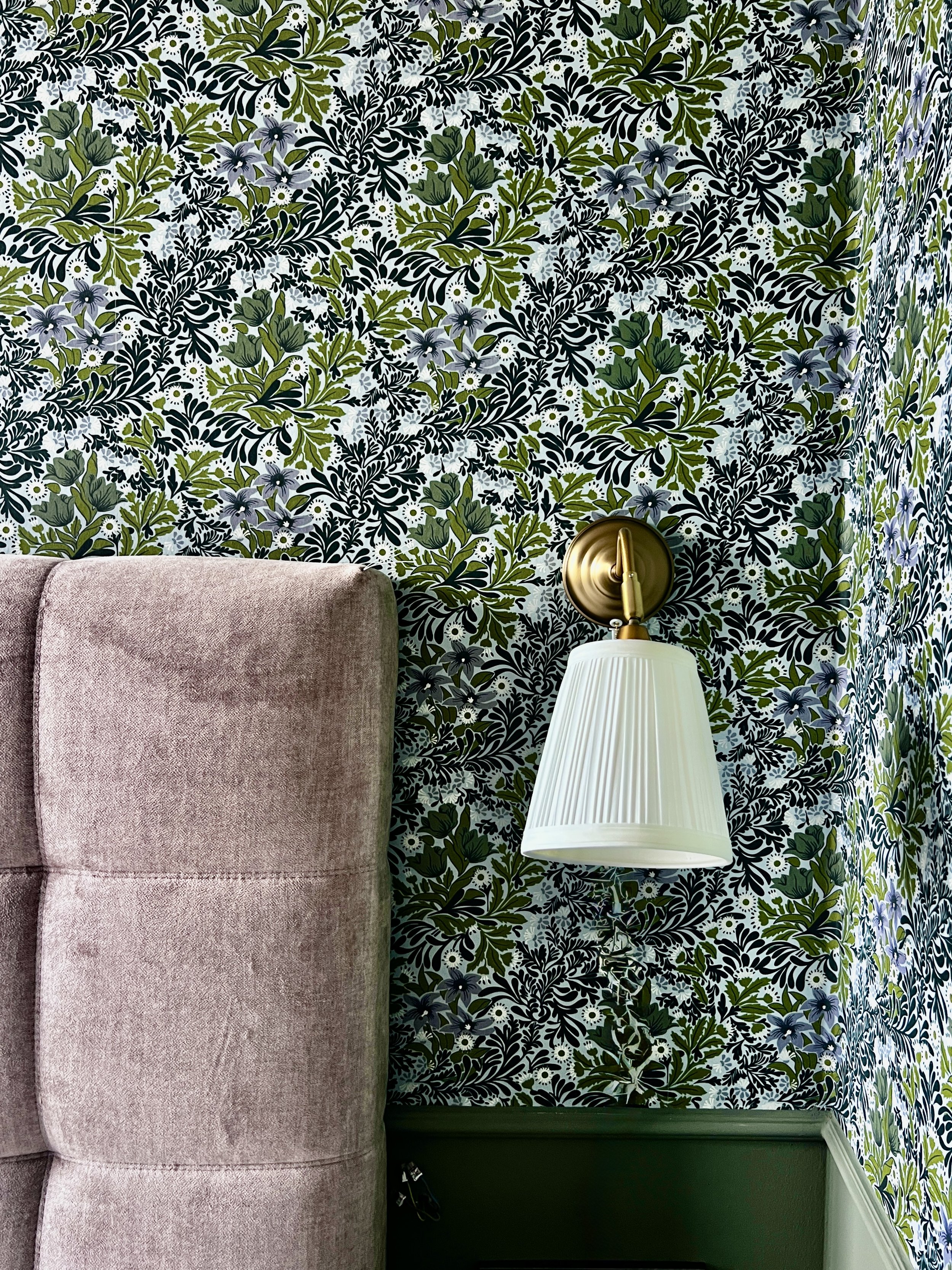 Ein pinkfarbenes gepolstertes Kopfteil eines Bettes vor einer Wand mit grünen, schwarzen, weißen und hellblauen Pflanzenmustern. An der Wand ist eine weiße Wandlampe mit goldener Halterung befestigt.