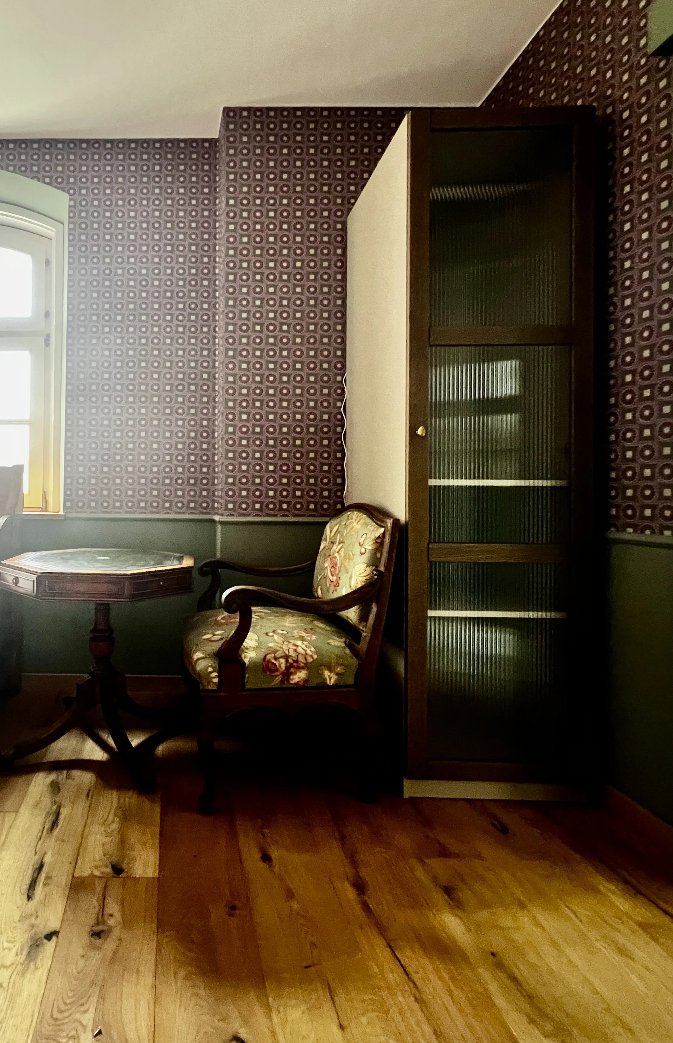 Ein Raum mit einem Fenster, einer gepolsterten antiken Holzarmlehnenstuhl mit Blumenmuster, einer runden Holztisch und einem hohen Regal mit Gläsern. Die Wände sind mit geometrisch gemusterter Tapete bekleidet, der Boden ist aus Holz.