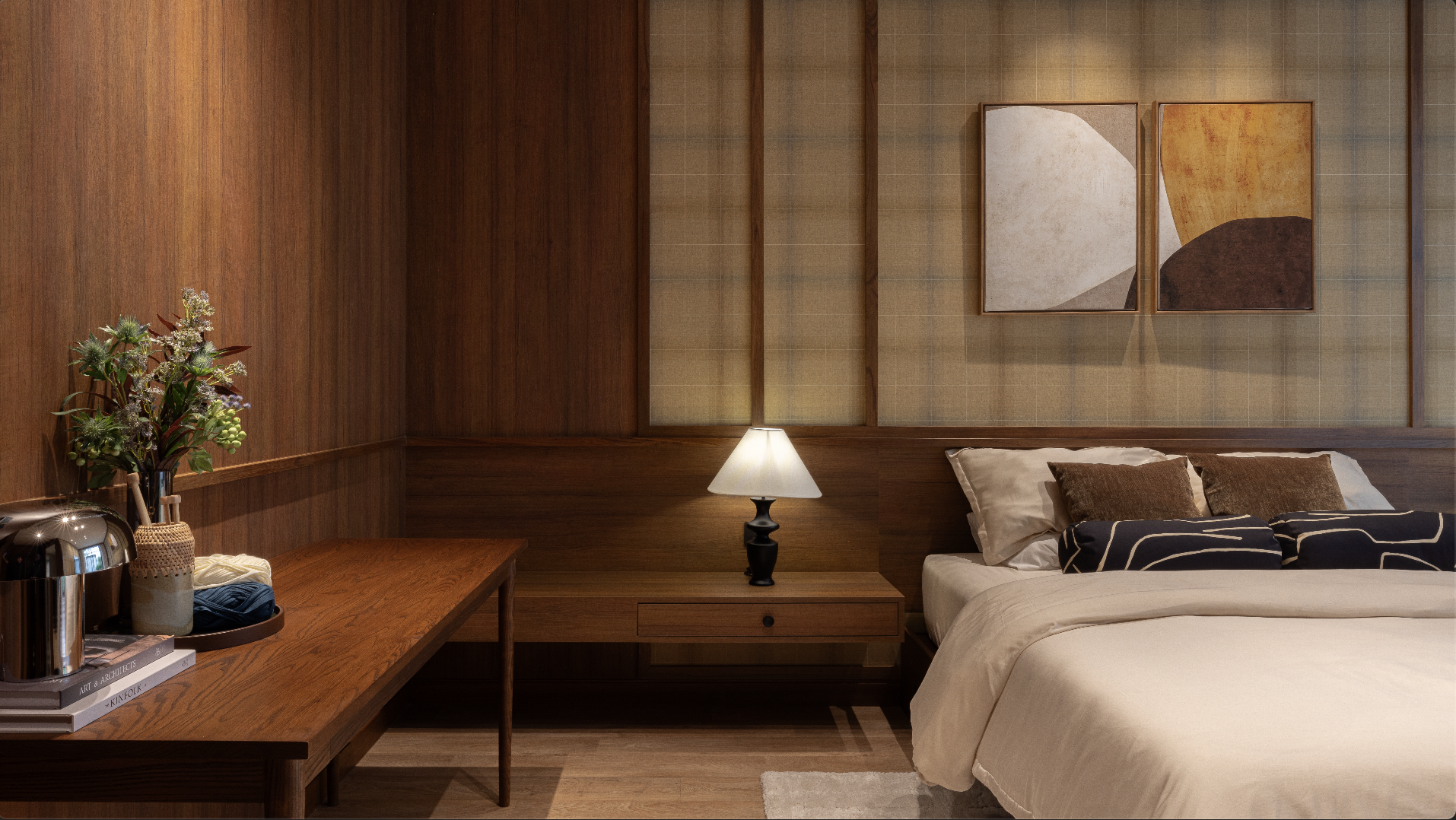 A bedroom with wooden wall paneling, a bed with white linens and brown pillows, a nightstand with a black table lamp, a wooden desk with a vase of flowers, books, and a silver water pitcher, and abstract art paintings on the wall.