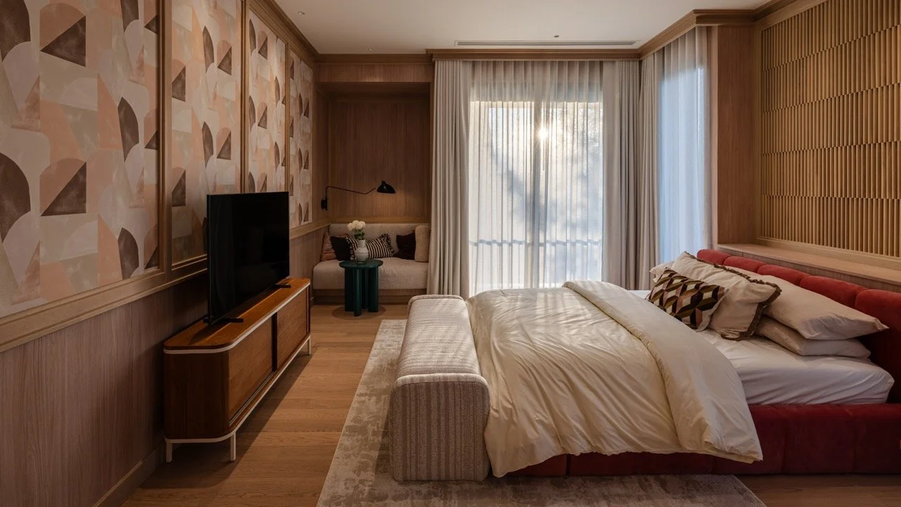 Modern bedroom with wooden paneled walls, a large bed with white bedding and patterned pillows, a TV on a wooden stand, a small sitting area with a couch, and large windows with sheer curtains letting in sunlight.