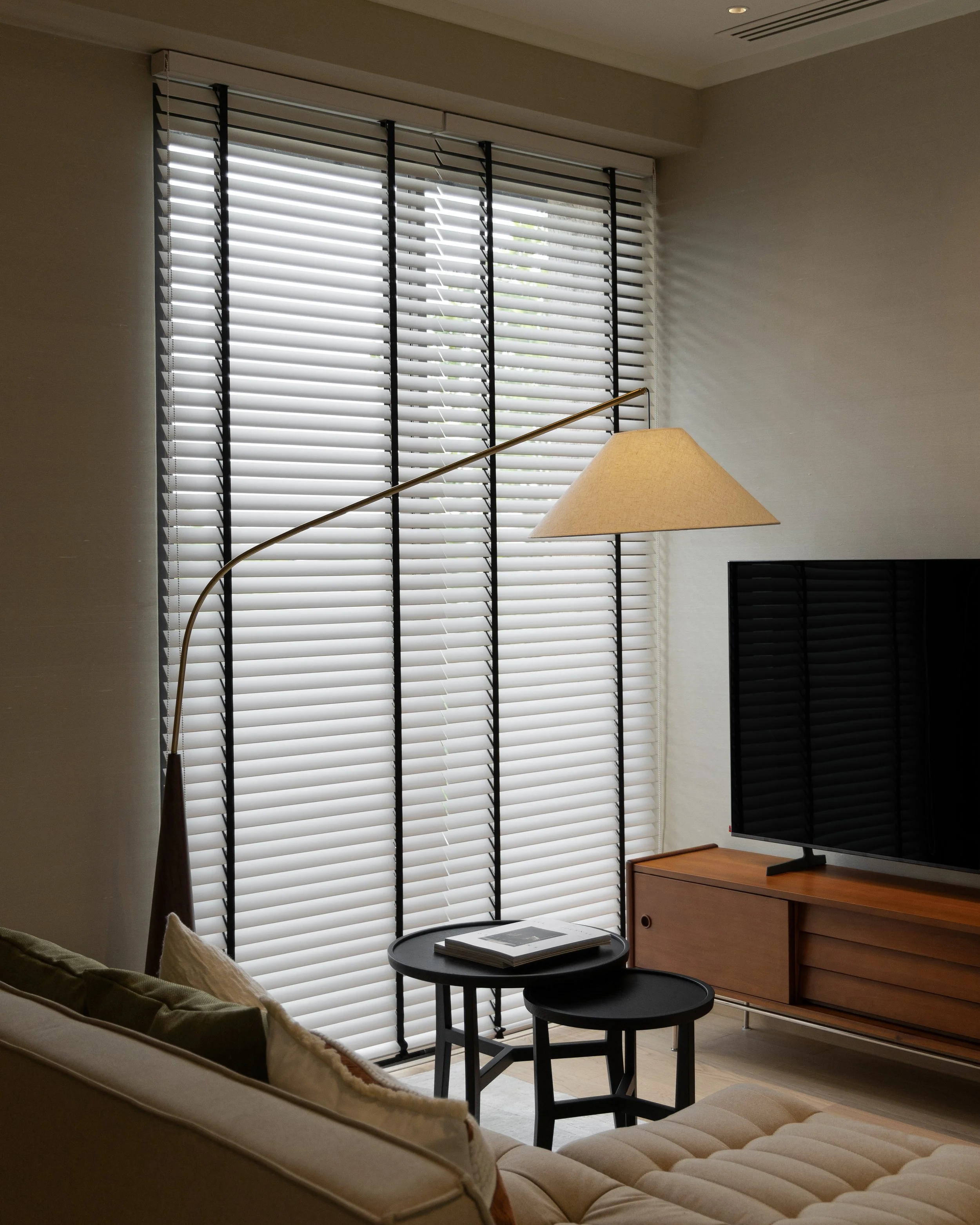 Living room with large window covered by horizontal blinds, beige sofa with pillows, modern floor lamp with beige shade, black side table with magazines, and wooden TV stand with flat-screen TV.
