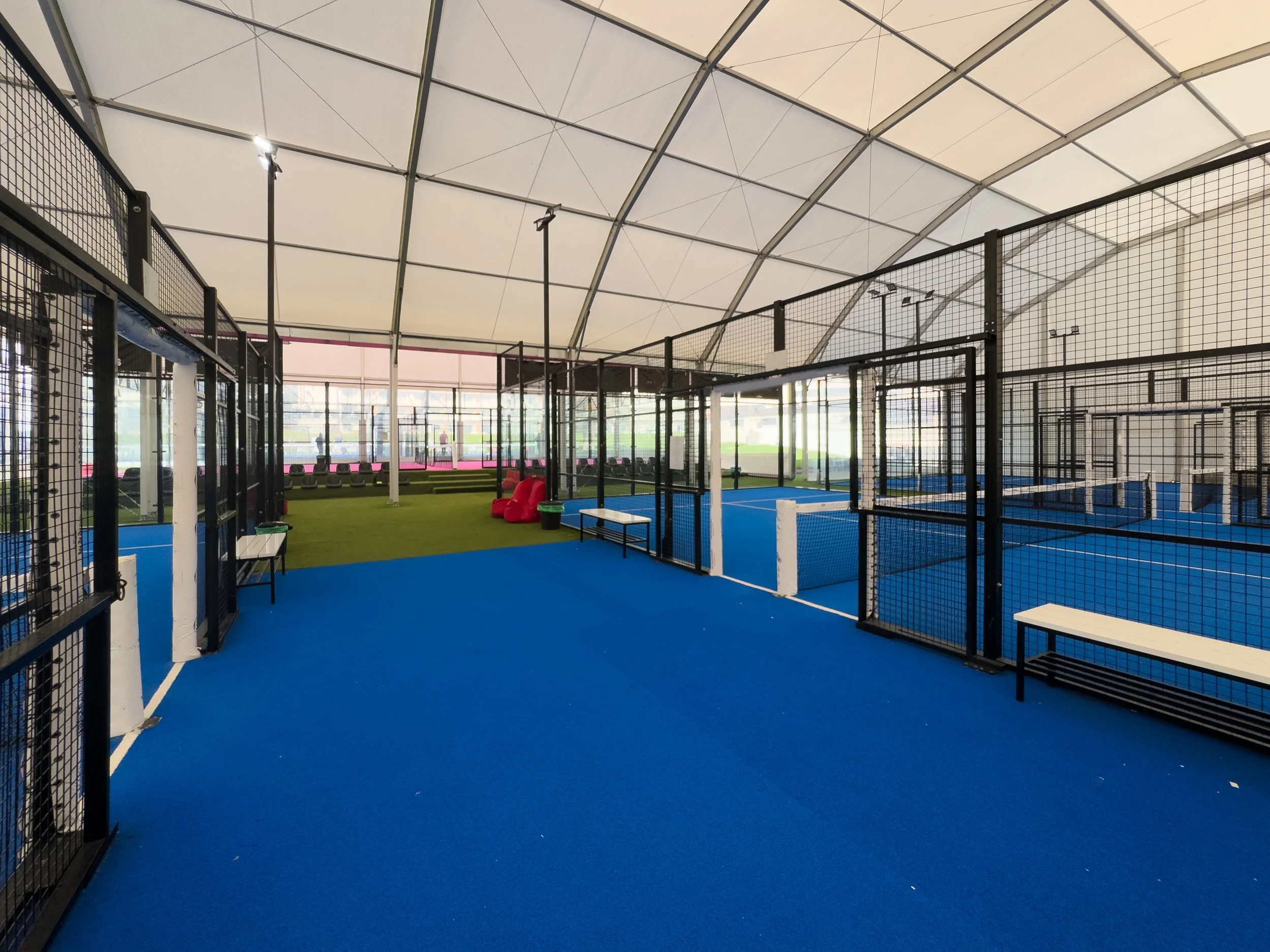 Innenansicht einer Padelhalle mit blauen Padelplätzen, einem grünen Bereich und Sitzgelegenheiten.