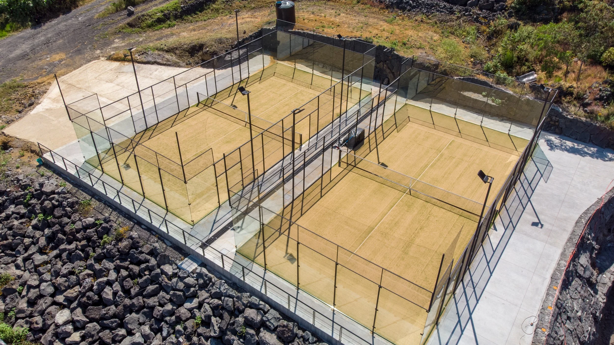 Luftaufnahme von vier Padelplätzen mit Sandbelag, umgeben von hohen Gittern und Beleuchtungsanlagen, in einer bergigen Landschaft.