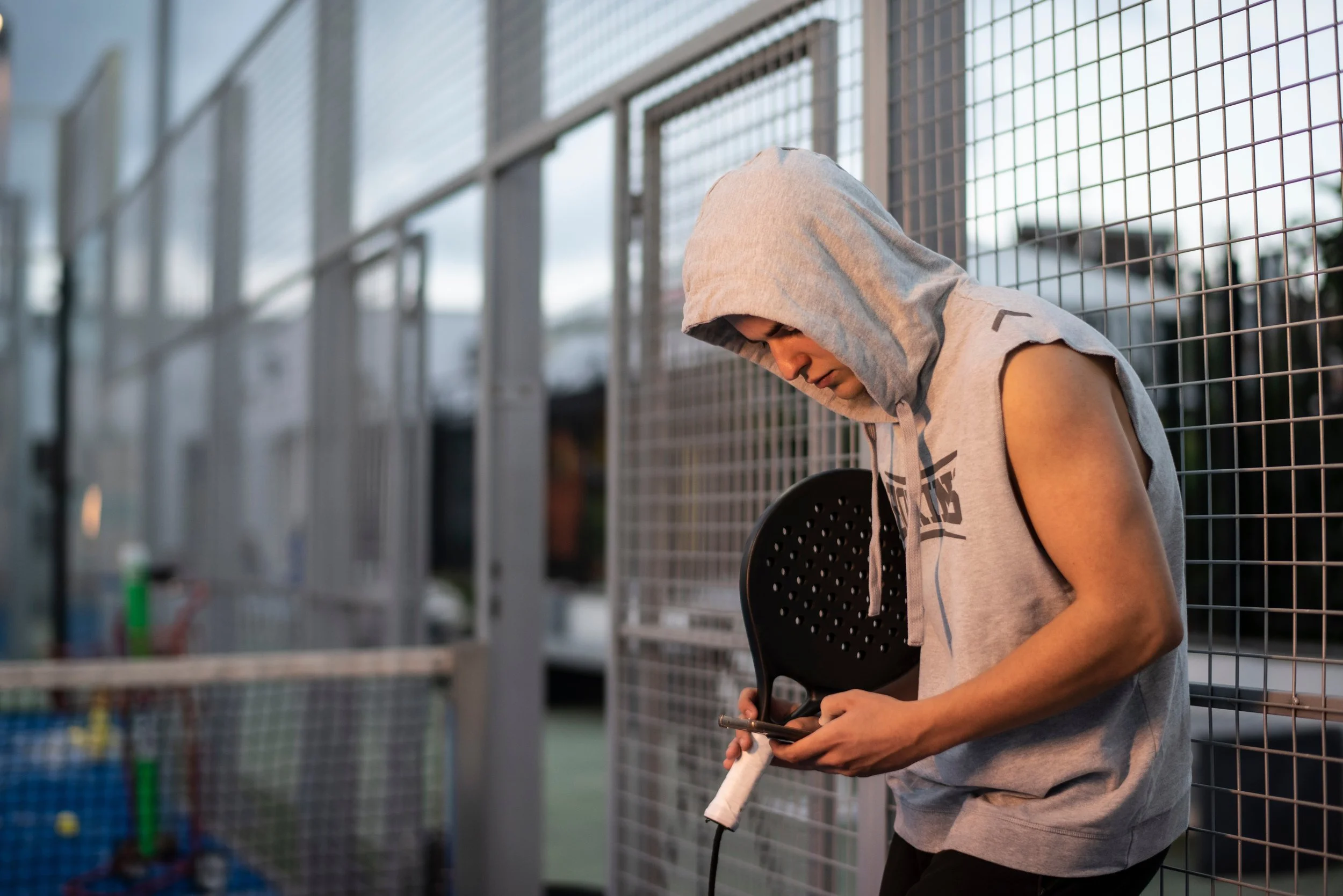 Junge Person mit Kapuzenpullover, der einen Padelhemd hält, steht vor einem Metallzaun auf einem Padelplatz, Blick nach unten, Abendlicht