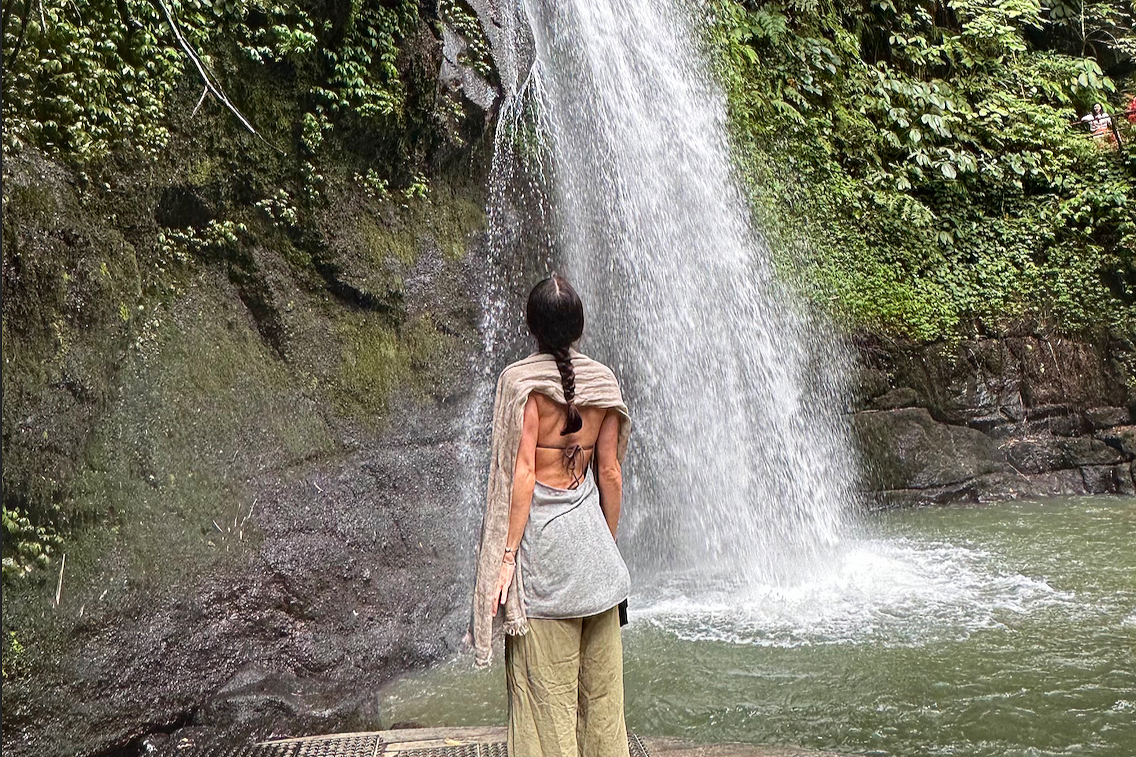 Person with long dark hair and braid, wearing a gray skirt and beige shawl, standing on a wooden platform, facing a waterfall in a natural setting.