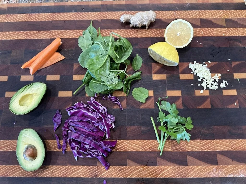 Assorted chopped vegetables and herbs on a wooden cutting board, including avocado, carrot, cilantro, purple cabbage, garlic, ginger, lemon, and spinach.