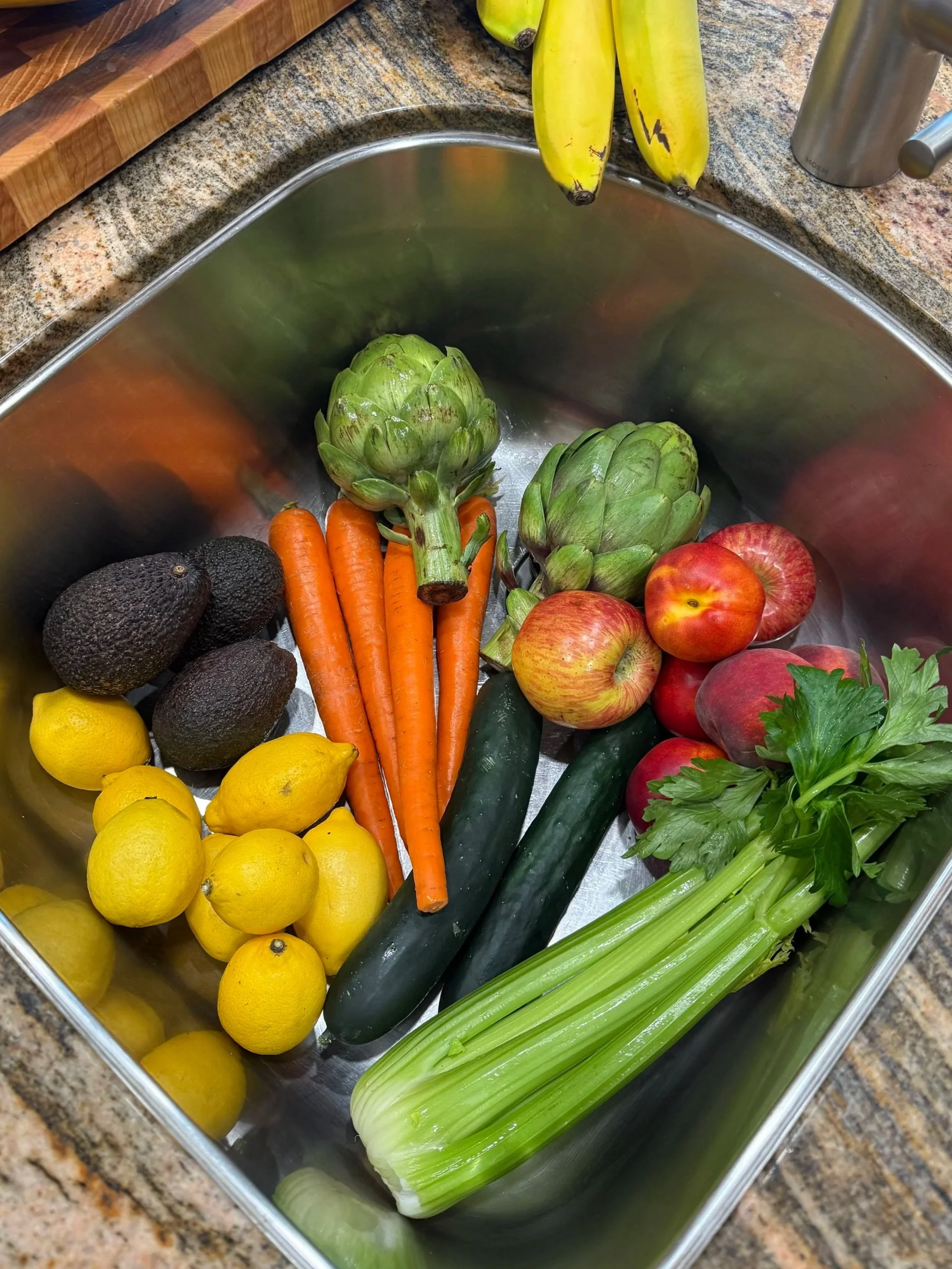 A kitchen sink filled with fresh vegetables and fruits, including lemons, avocados, carrots, artichokes, cucumbers, apples, celery, and peaches.