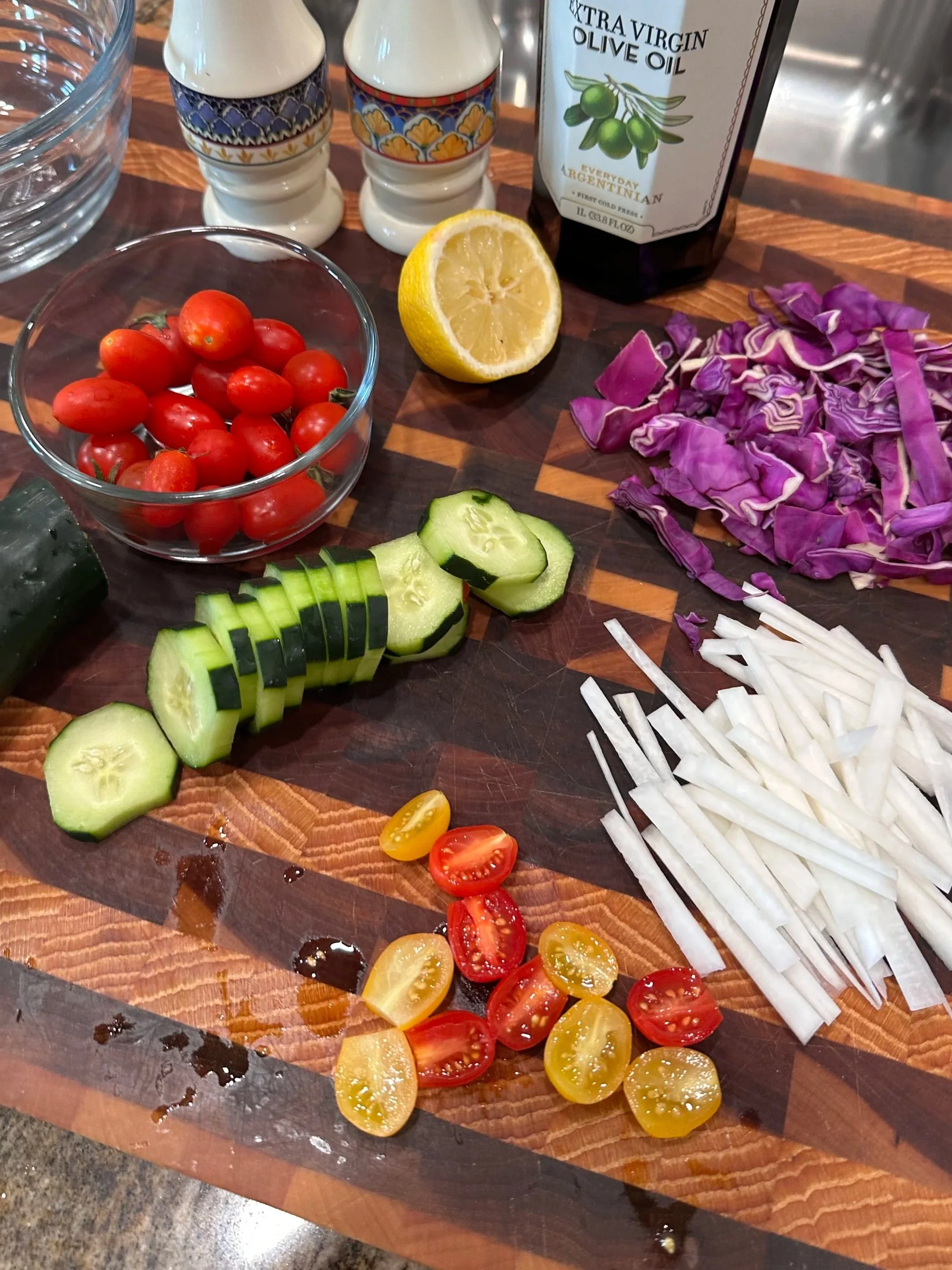 An assortment of chopped vegetables and ingredients on a wooden cutting board, including cherry tomatoes, cucumber slices, shredded purple cabbage, sliced white onion, and halved yellow and red cherry tomatoes. A halved lemon, a bottle of extra virgin olive oil, and salt and pepper shakers are also visible on the countertop.