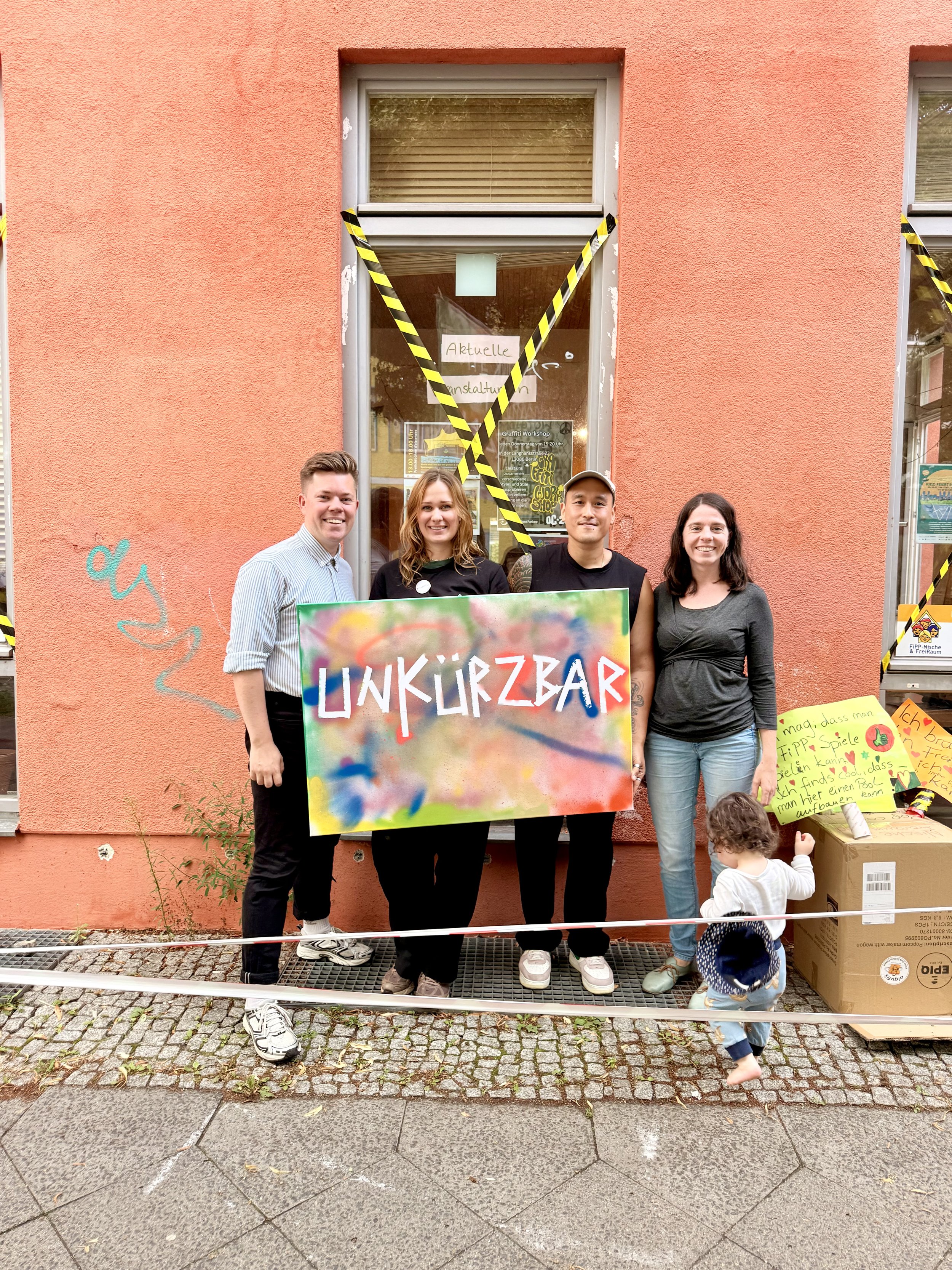 Vier Menschen stehen vor einem pinkfarbenen Gebäude, halten ein bunt bemaltes Schild mit der Aufschrift "INKÜRZAR". Ein Kind steht vor ihnen und schaut auf ein großes Pappkarton. Im Hintergrund sind Bauzäune mit schwarz-gelben Warnbändern und ein Fenster mit Notizzetteln.