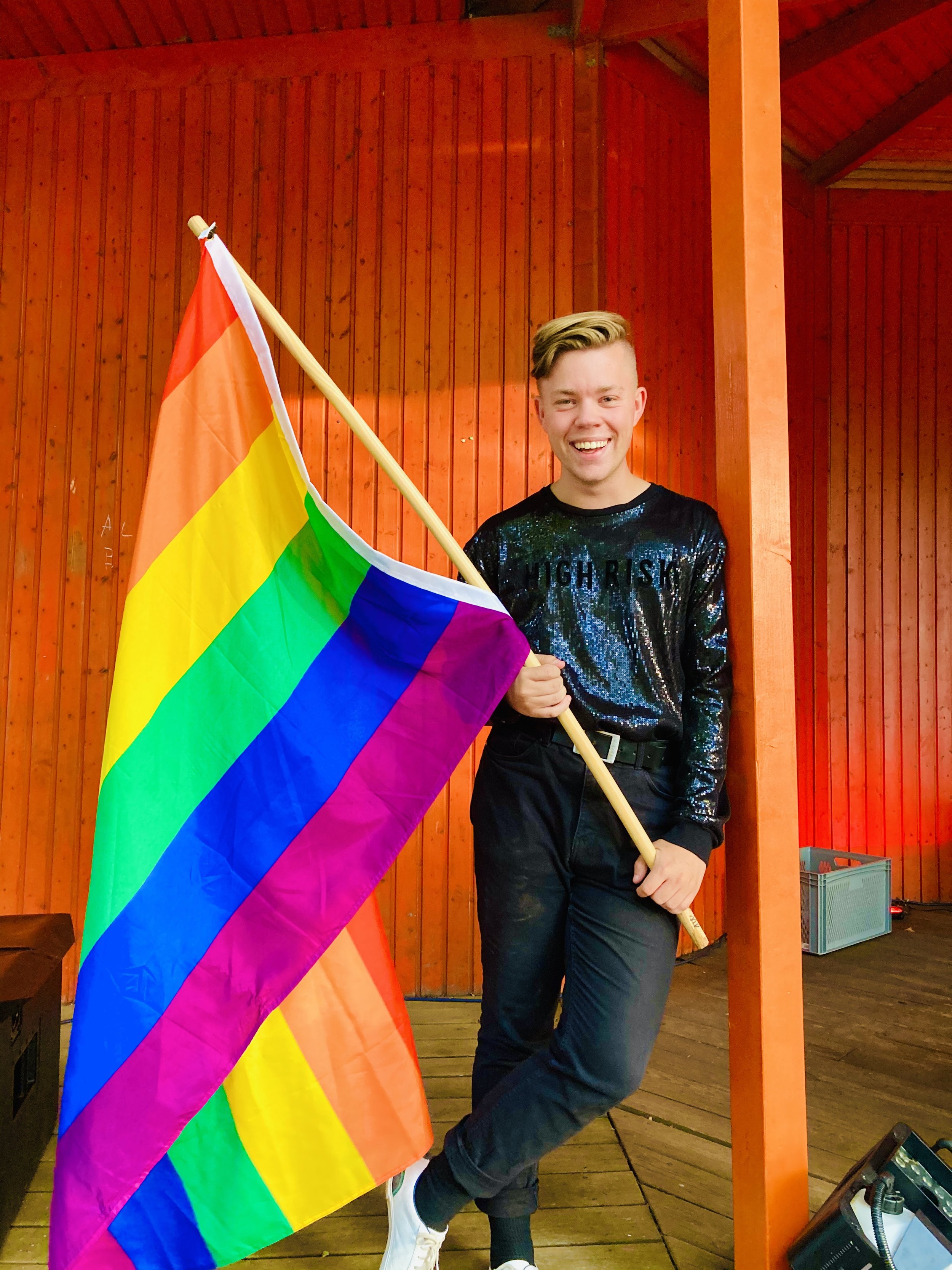 Ein lachender junger Mensch hält eine Regenbogenfahne an einem Holzmast und steht vor einer roten Holzwand.