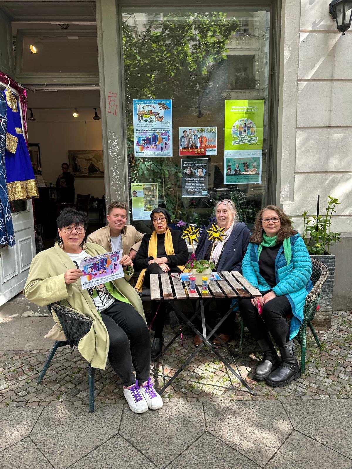 Fünf Menschen sitzen bequem an einem kleinen Tisch vor einem Café, einige halten Flyer für eine Veranstaltung. Die Gruppe besteht aus Frauen und einem Mann, alle lächelnd. Es sind bunte Dekorationen auf dem Tisch, darunter zwei Windmühlen mit Bienenmotiven.