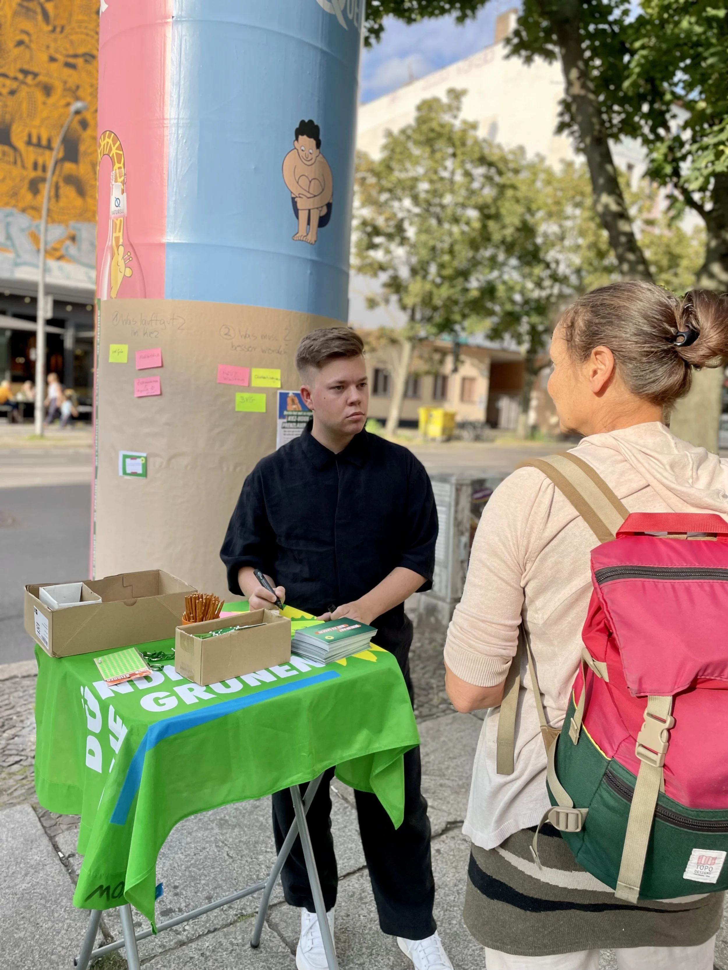 Ein Junge steht an einem Stand mit einer grünen Tafel, während eine älteren Frau mit einem roten Rucksack mit ihm spricht. Es befinden sich Post-its und Broschüren auf dem Tisch.