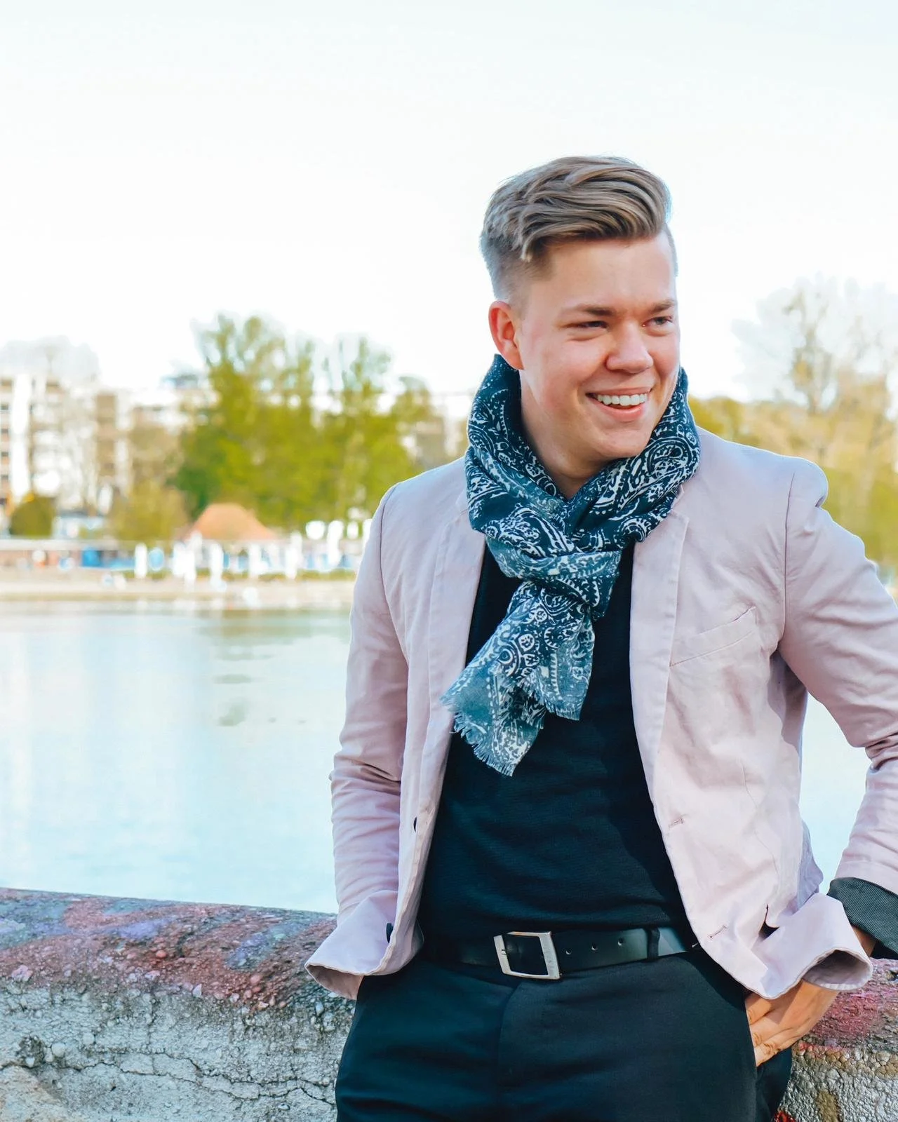 Ein lachender junger Mann in Freizeitkleidung steht an einem Fluss und schaut in die Ferne, im Hintergrund Bäume und Gebäude.